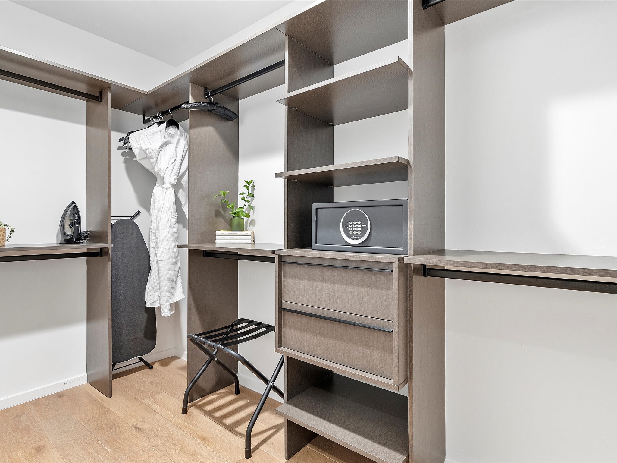 A spacious walk-in closet features dark wood shelving and hanging rods, with a white bathrobe, ironing board, and a small safe visible, all set against white walls and light wood flooring.