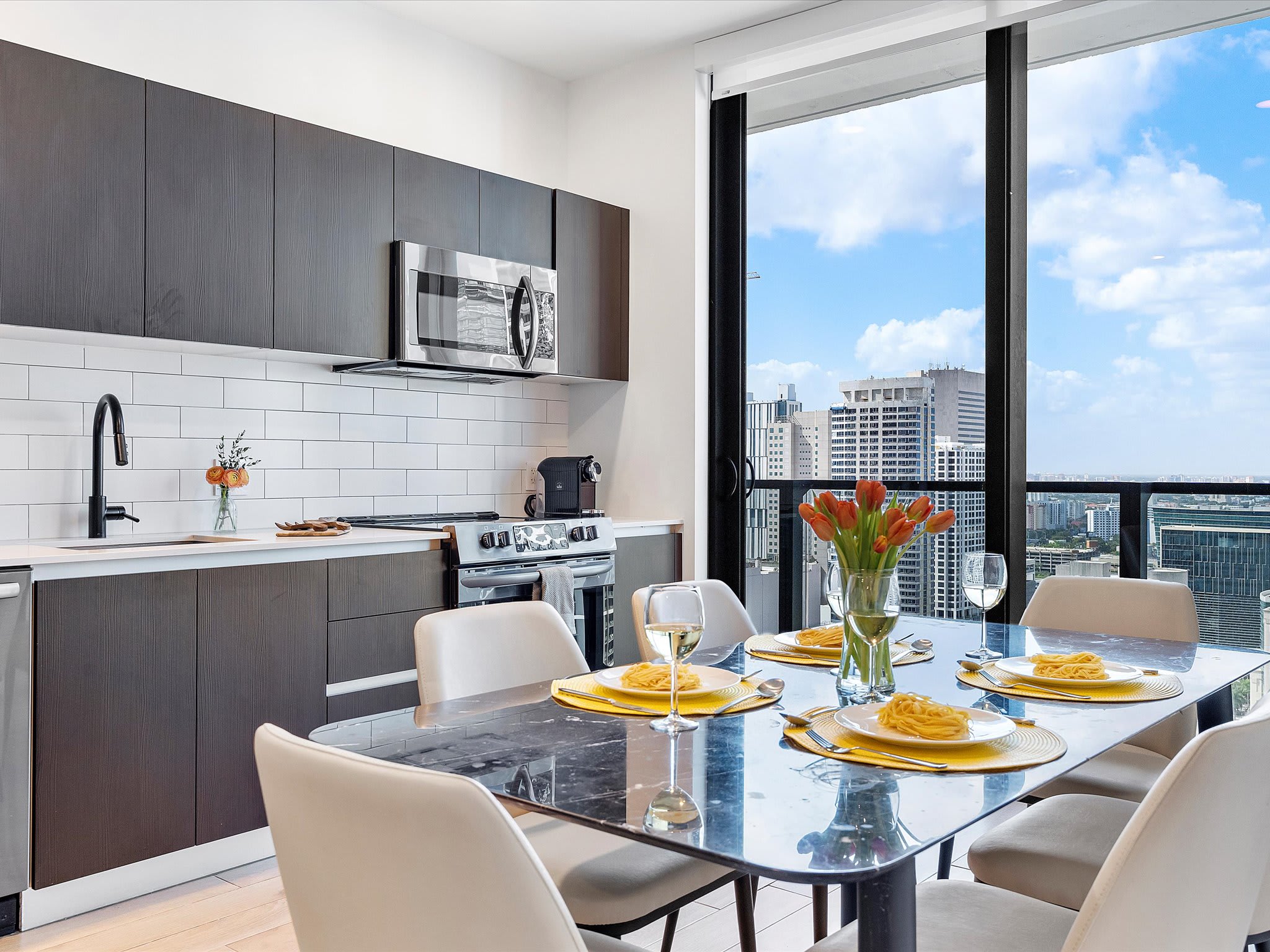 Modern kitchen with dark wood cabinets, white subway tile backsplash, stainless steel appliances, and a black faucet, adjacent to a dining area with a black marble table set for four, offering expansive city views through floor-to-ceiling windows.