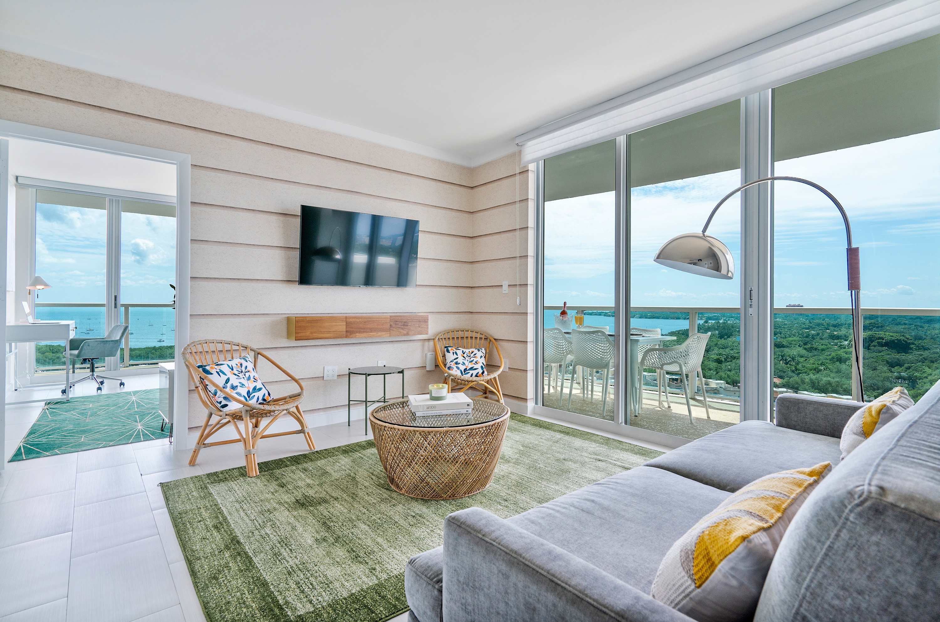 Cozy living room with a gray sofa, two wicker chairs, and a round wicker coffee table on a green area rug. Floor-to-ceiling sliding glass doors lead to a balcony with bay views, and a wall-mounted TV is visible.