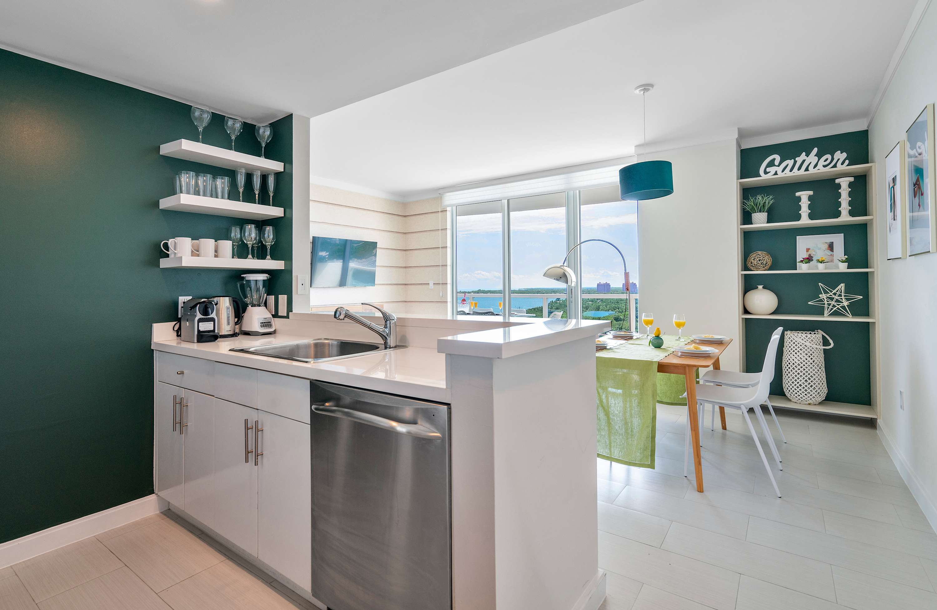Modern kitchen with white cabinetry, stainless steel appliances, and a dark teal accent wall with floating shelves displaying glassware. A breakfast bar separates the kitchen from the dining area, which features a wooden table and white chairs.