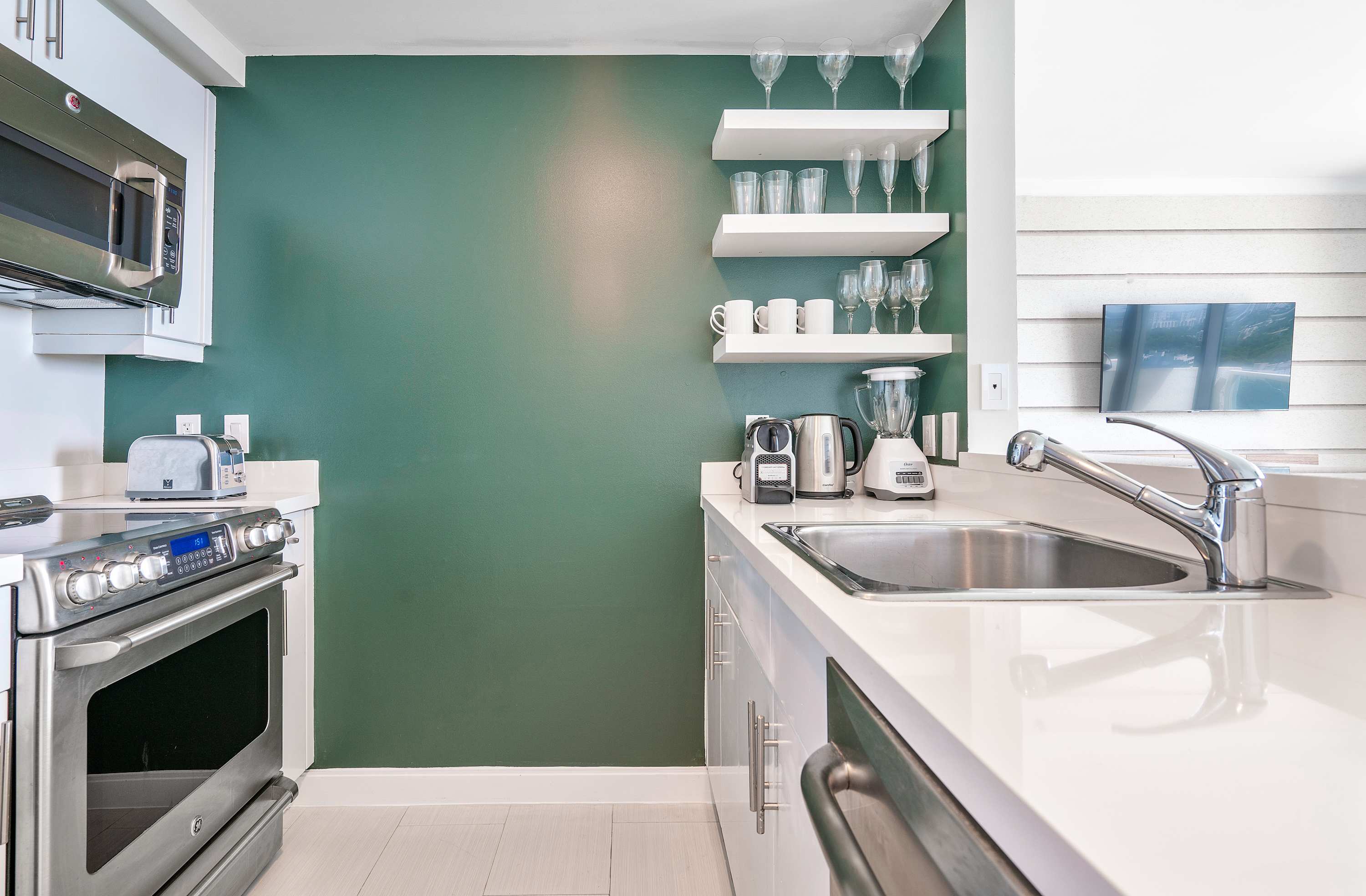 Modern kitchen with white countertops, stainless steel appliances including a microwave, oven, and sink, and a vibrant teal accent wall with white floating shelves displaying glassware.