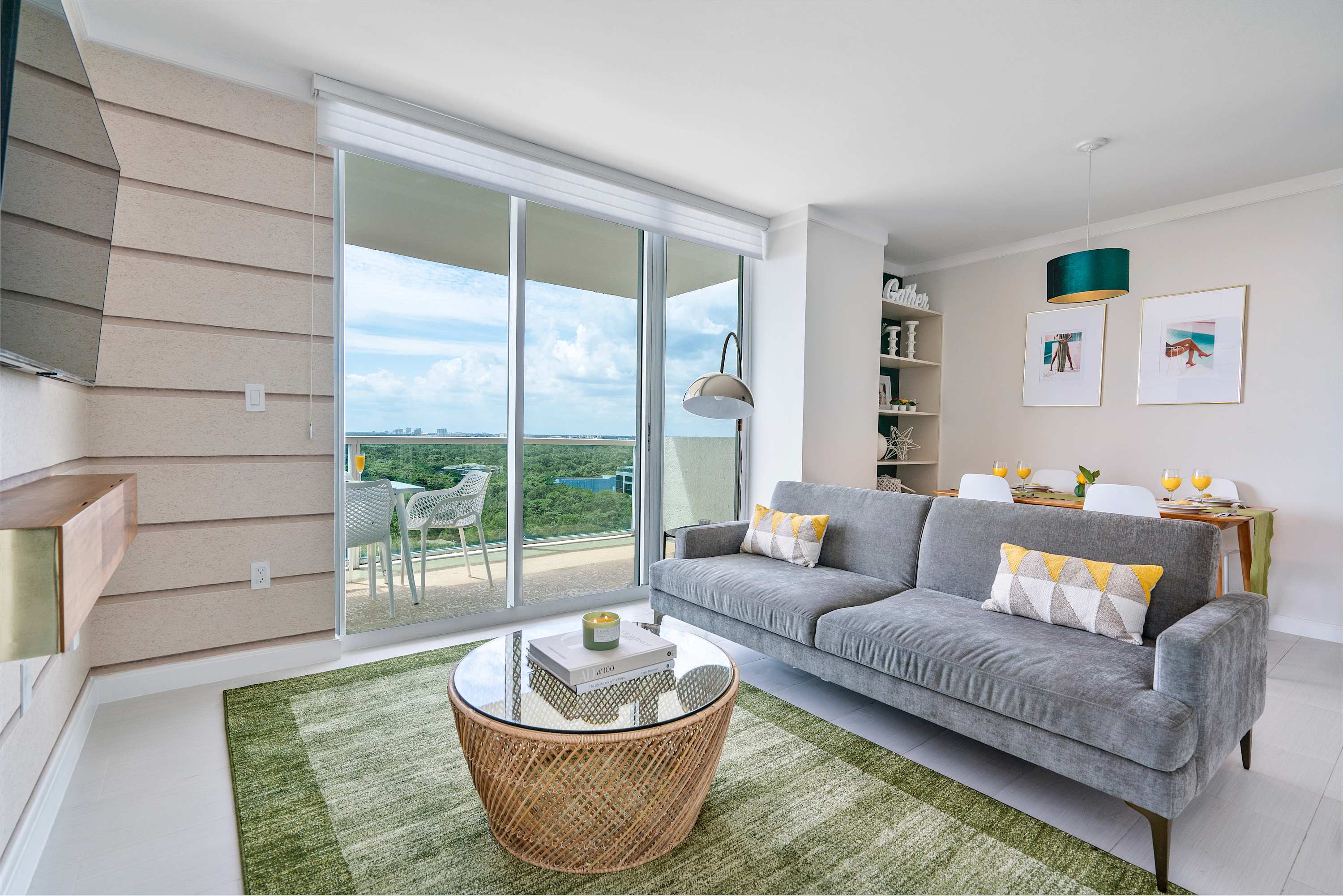 Bright living room featuring a comfortable gray sofa, a round glass-top coffee table, and a green area rug. Floor-to-ceiling windows provide ample natural light and views of the bay and surrounding greenery.