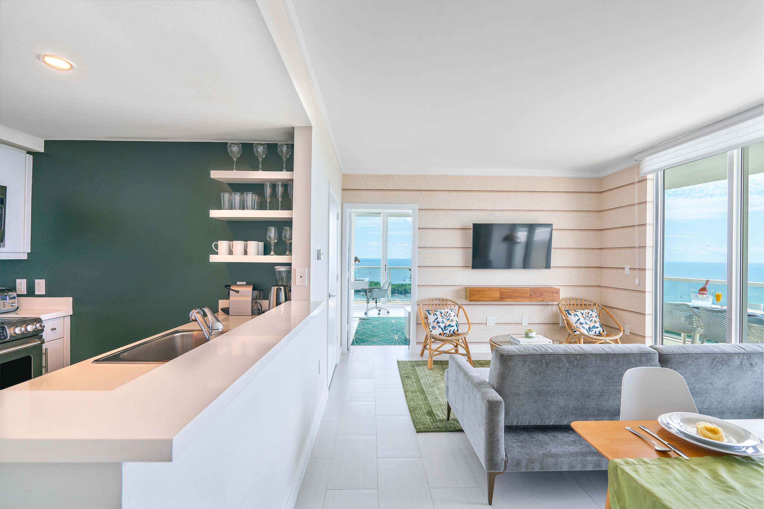 Open-concept view of the kitchen and living area, showing white kitchen cabinets and a breakfast bar on the left, leading into the living room with a gray sofa and a dining table. Floor-to-ceiling windows offer a glimpse of the bay view.