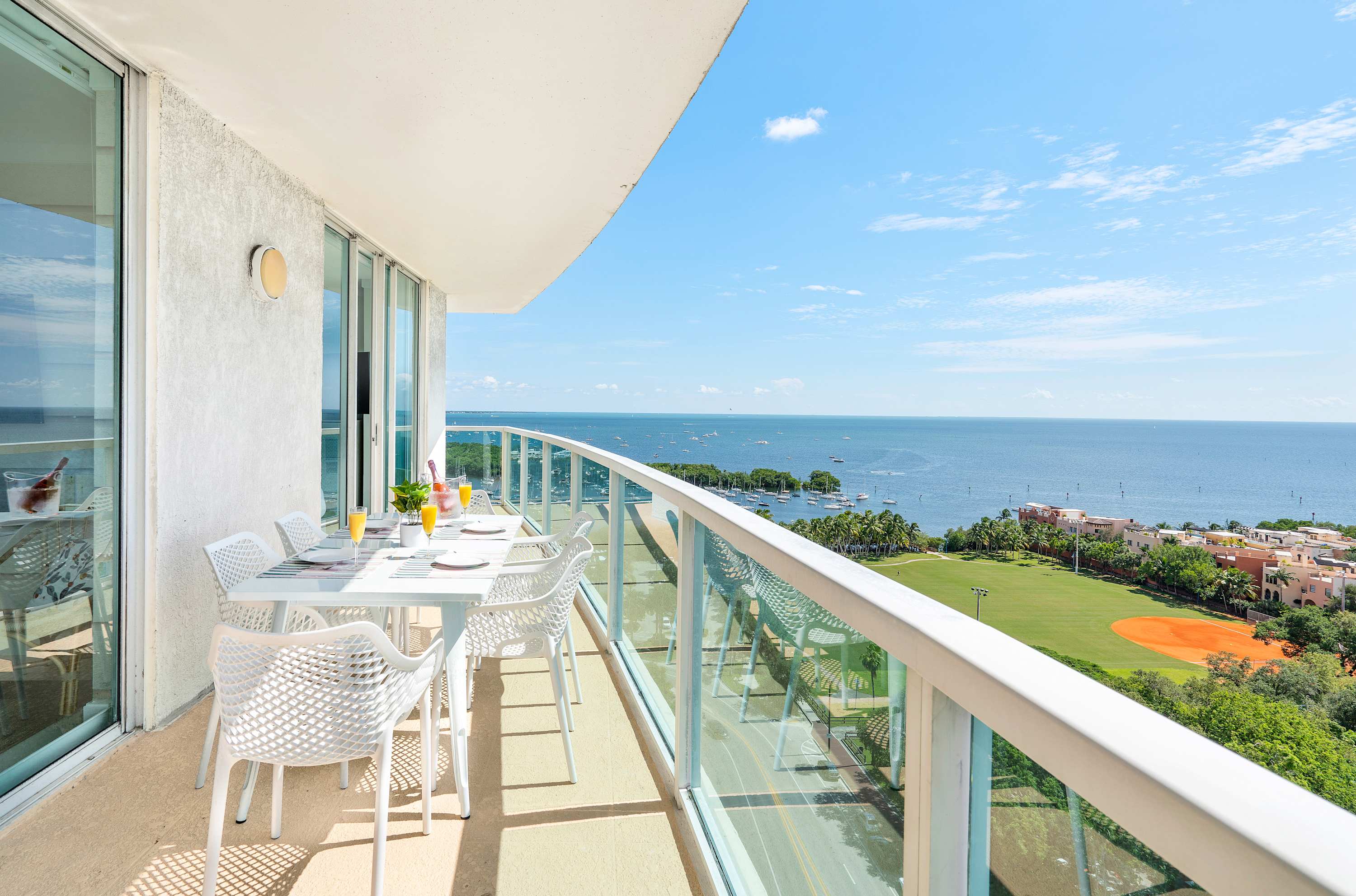 Spacious private balcony with a white outdoor dining set for six, offering panoramic views of the ocean, a marina with boats, and a green park with a baseball field.