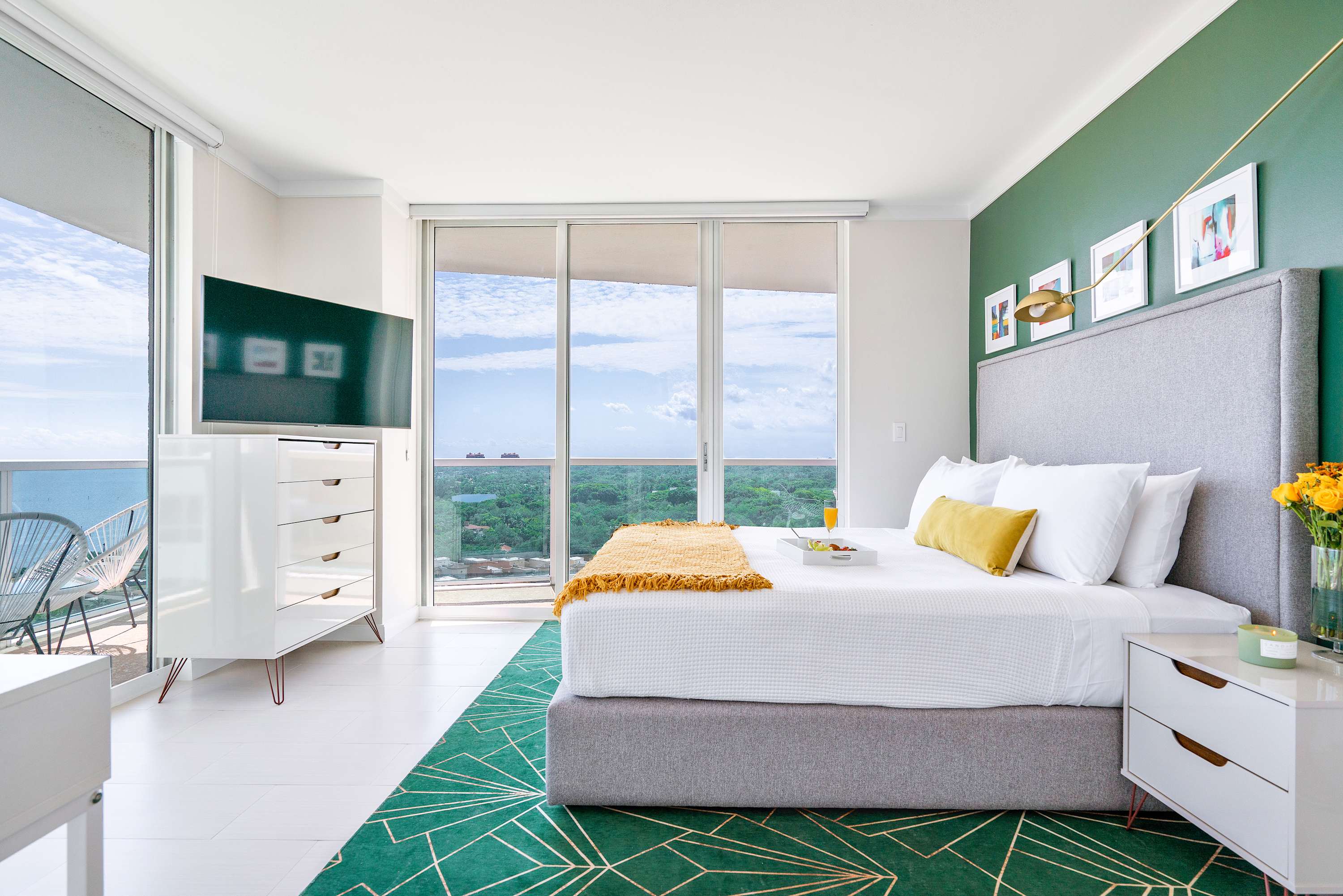 Modern bedroom featuring a large bed with a gray upholstered headboard against a dark teal accent wall, complemented by a vibrant green geometric rug. Floor-to-ceiling windows provide abundant natural light and views of the surrounding landscape.