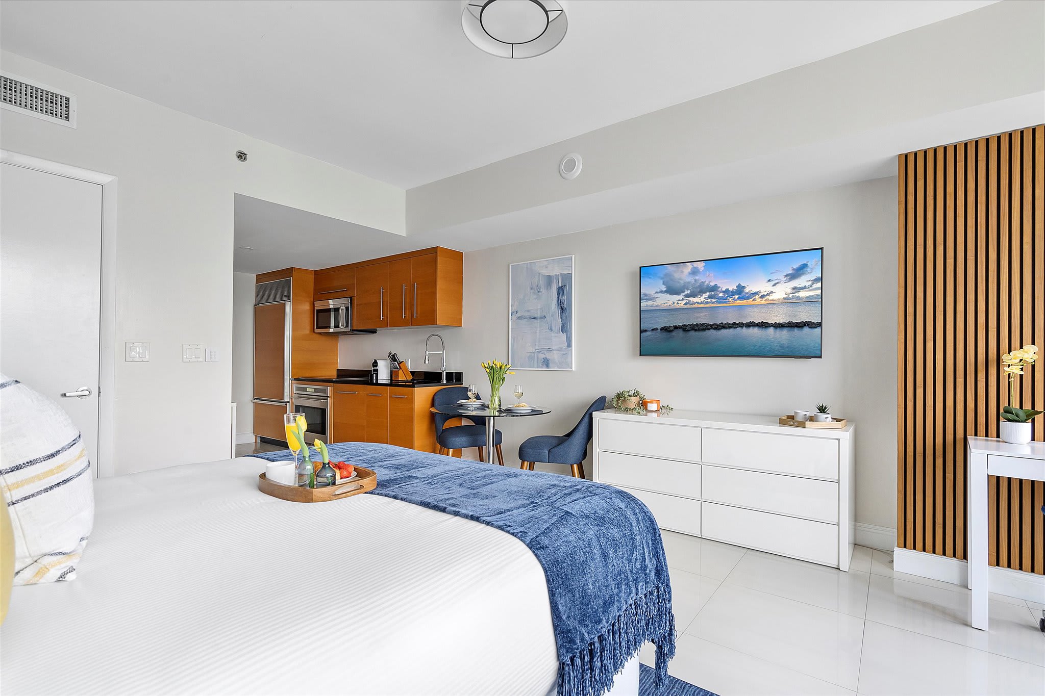 Open-concept studio apartment showcasing a queen bed, a compact kitchenette with light wood cabinetry and stainless steel appliances, and a small dining area for two. A wall-mounted flat-screen TV and a vertical slatted wood accent wall add modern touches.