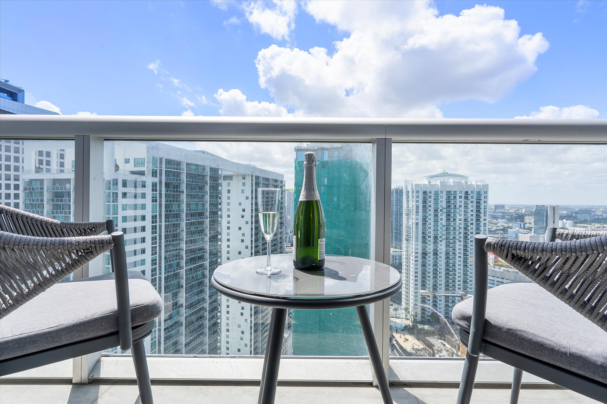 Private outdoor balcony furnished with two grey woven chairs and a small round table, offering expansive city views of numerous high-rise buildings. A bottle of champagne and a flute are set on the table, suggesting a relaxing experience.