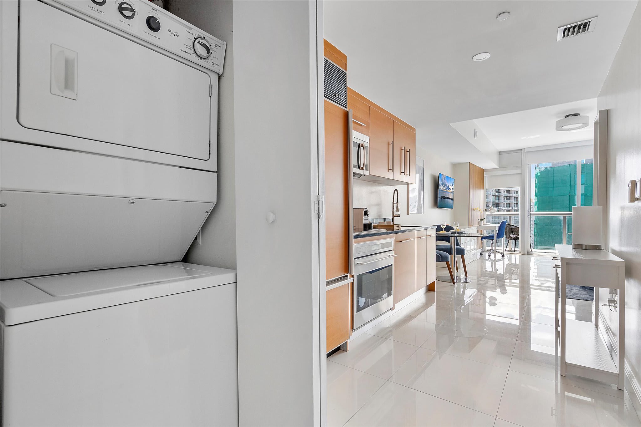 A compact laundry area featuring a stacked white washer and dryer unit, neatly integrated into a closet space. The adjacent open-concept area reveals a modern kitchen with light wood cabinets and a glimpse of the living space.