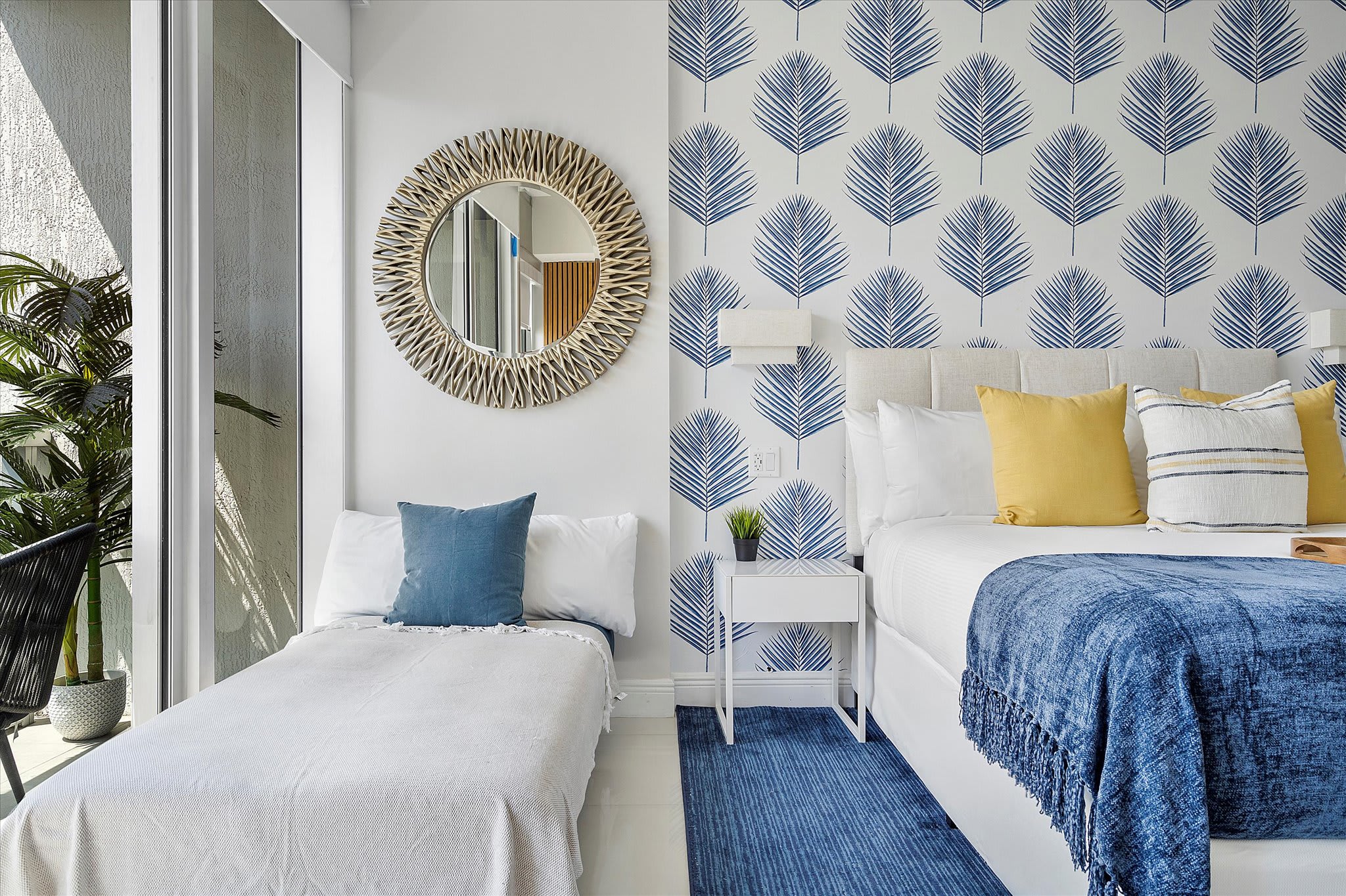 Contemporary studio bedroom featuring a queen bed and an additional twin daybed, both dressed in crisp white linens. The room is brightened by a blue palm leaf wallpaper accent wall and a large round mirror, with access to a private balcony.