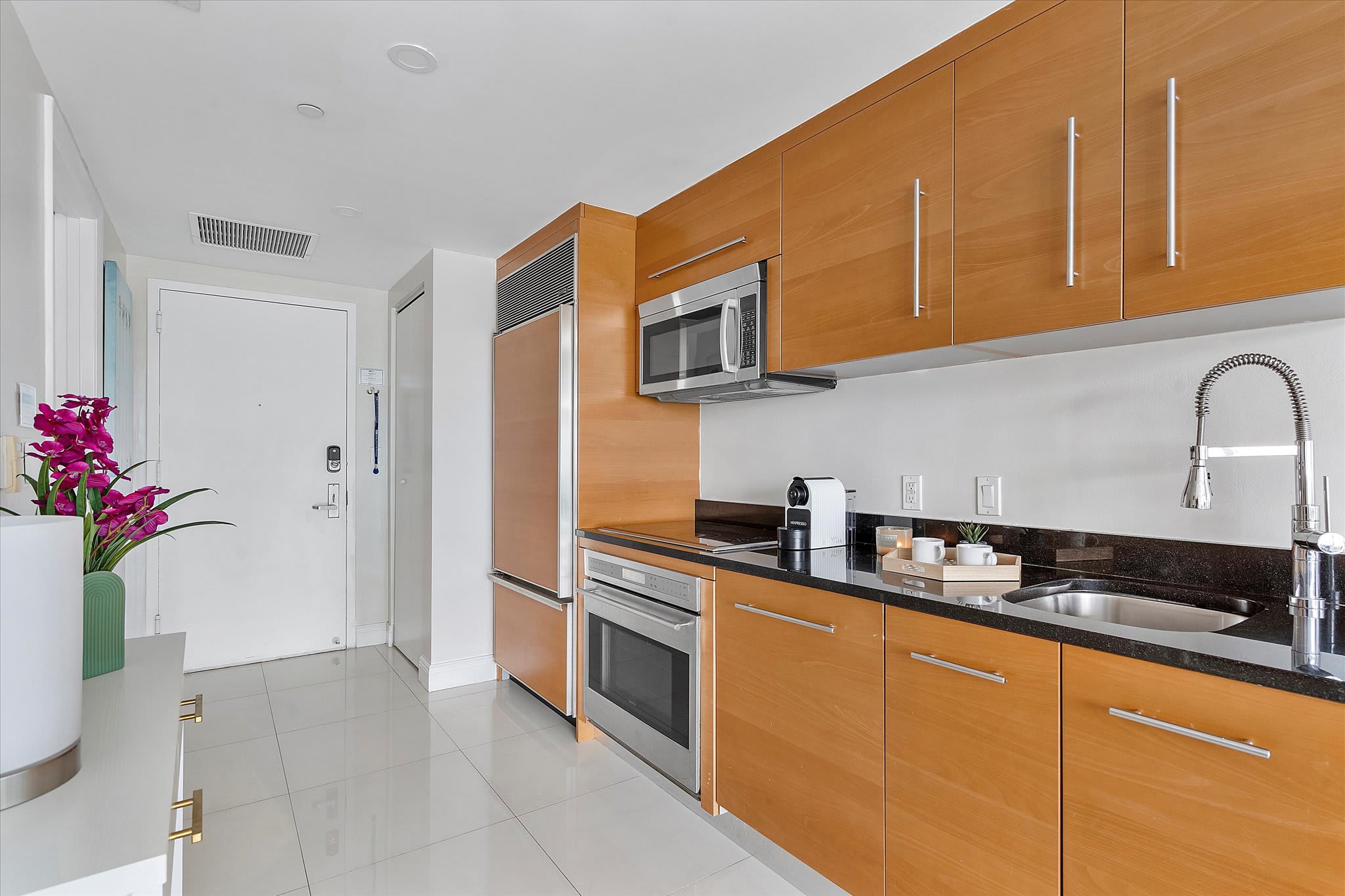 A well-equipped kitchen features light wood cabinetry, black granite countertops, and stainless steel appliances, including a refrigerator, microwave, and oven. A white tiled floor leads to the apartment entrance door.