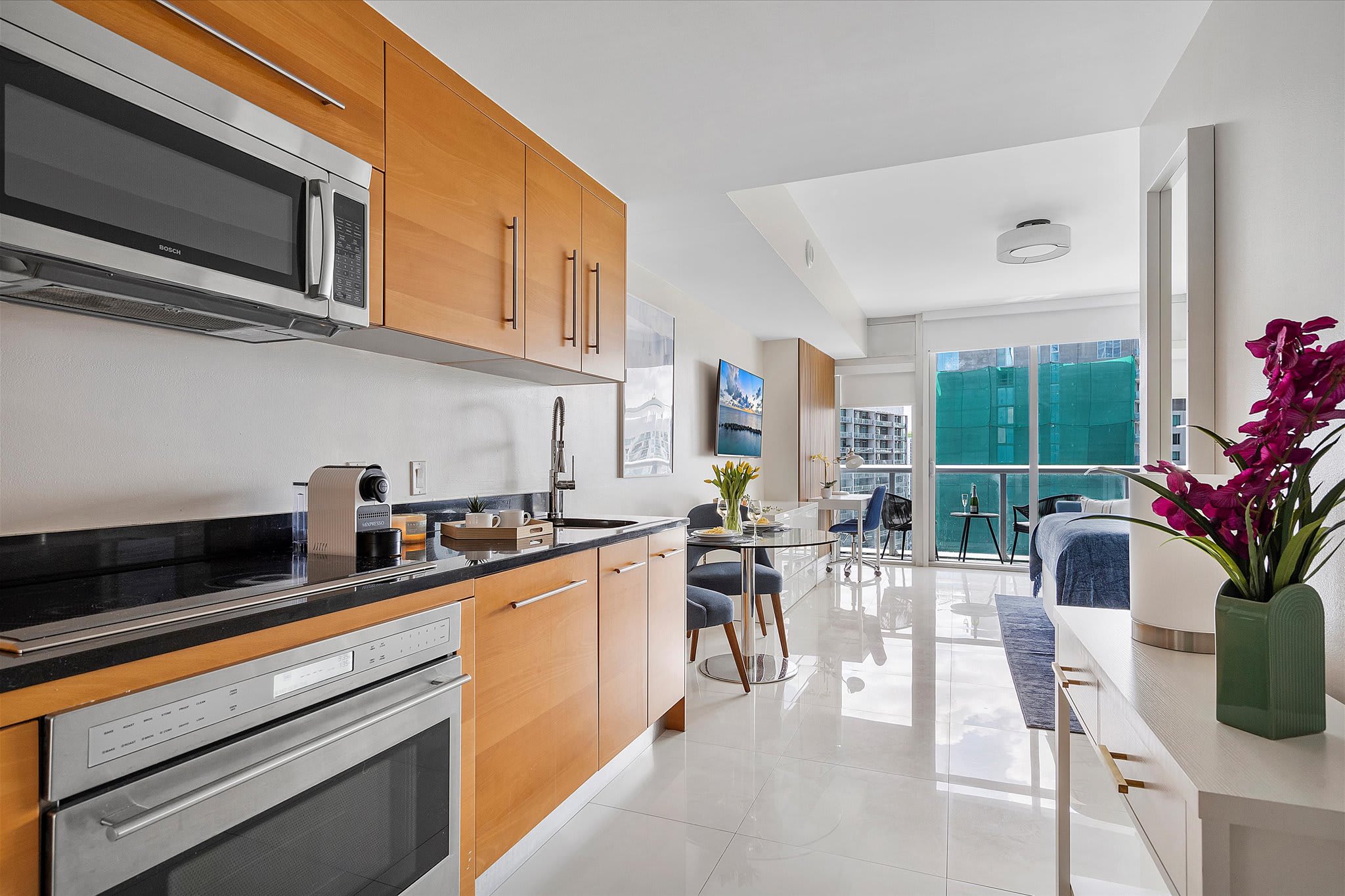 The modern kitchen with light wood cabinets and black countertops opens into a dining area with a round glass table and blue chairs, leading to a balcony with city views. White tiled floors reflect the bright natural light.