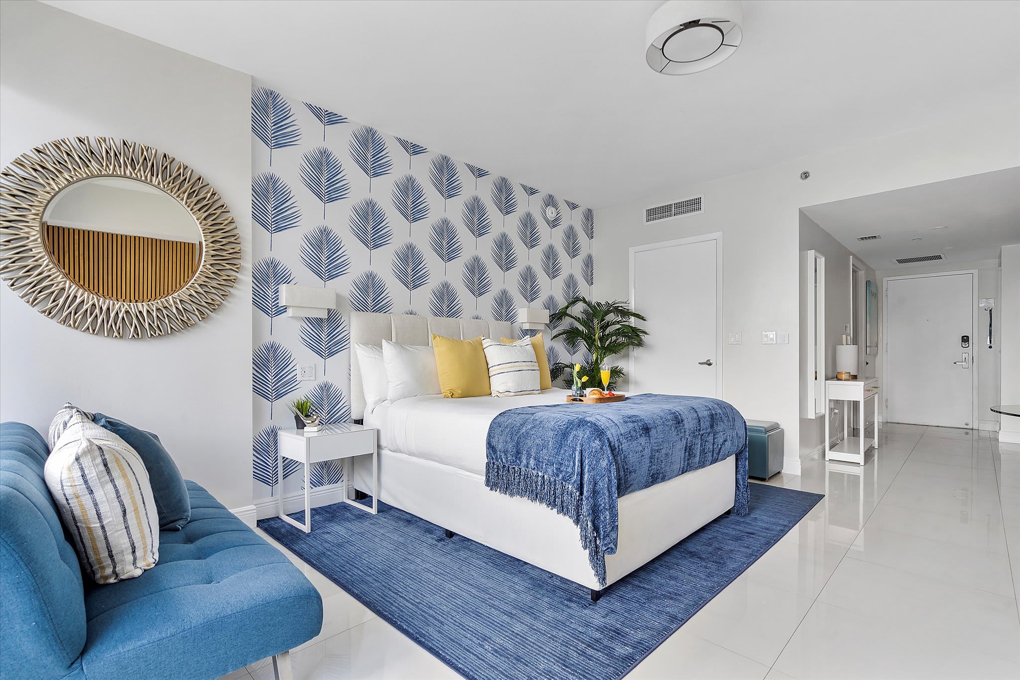 Bright and modern studio bedroom featuring a queen-sized bed with a white upholstered headboard, blue and yellow bedding, and a striking blue palm leaf wallpaper accent wall. A small blue sofa and a large round mirror with a textured frame complete the tropical-inspired decor.