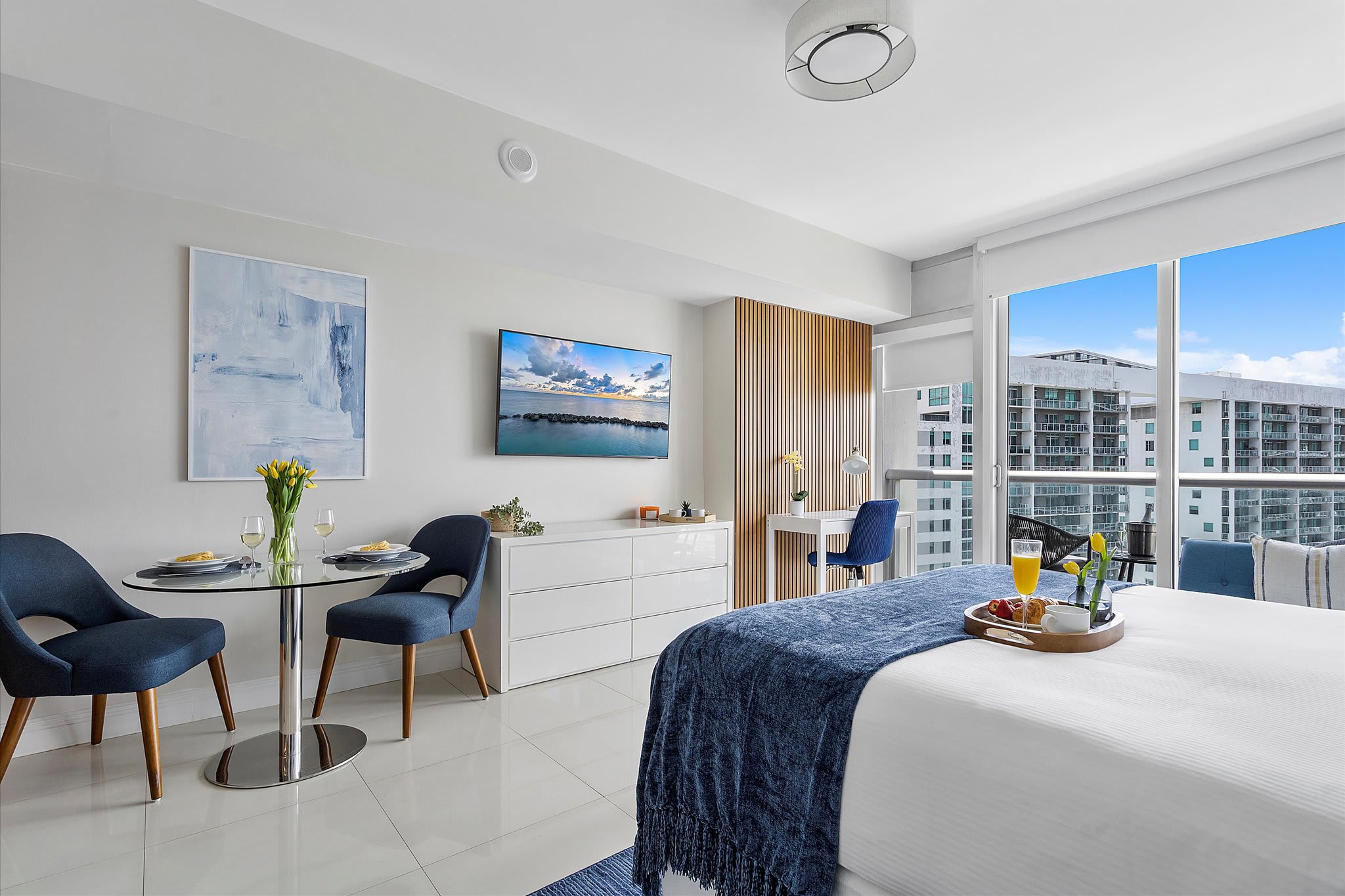 A bright studio apartment showcases a white bed with a blue throw, a small glass dining table with blue chairs, and a dedicated desk area. White tiled floors and a wall-mounted TV complete the modern, open-plan living space.