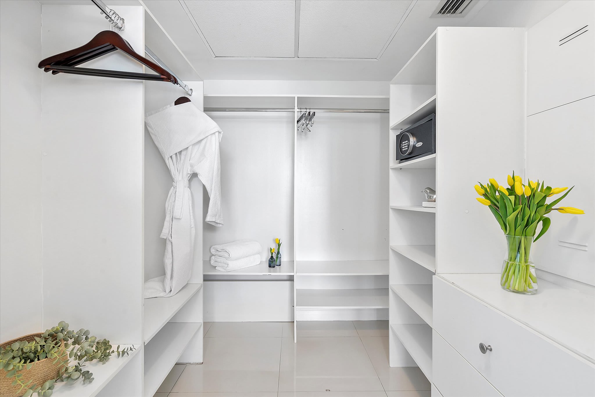 Spacious, all-white walk-in closet equipped with built-in shelving, drawers, and hanging rods, providing ample storage. A small safe is integrated into one of the shelves, and a white bathrobe hangs, indicating guest amenities.