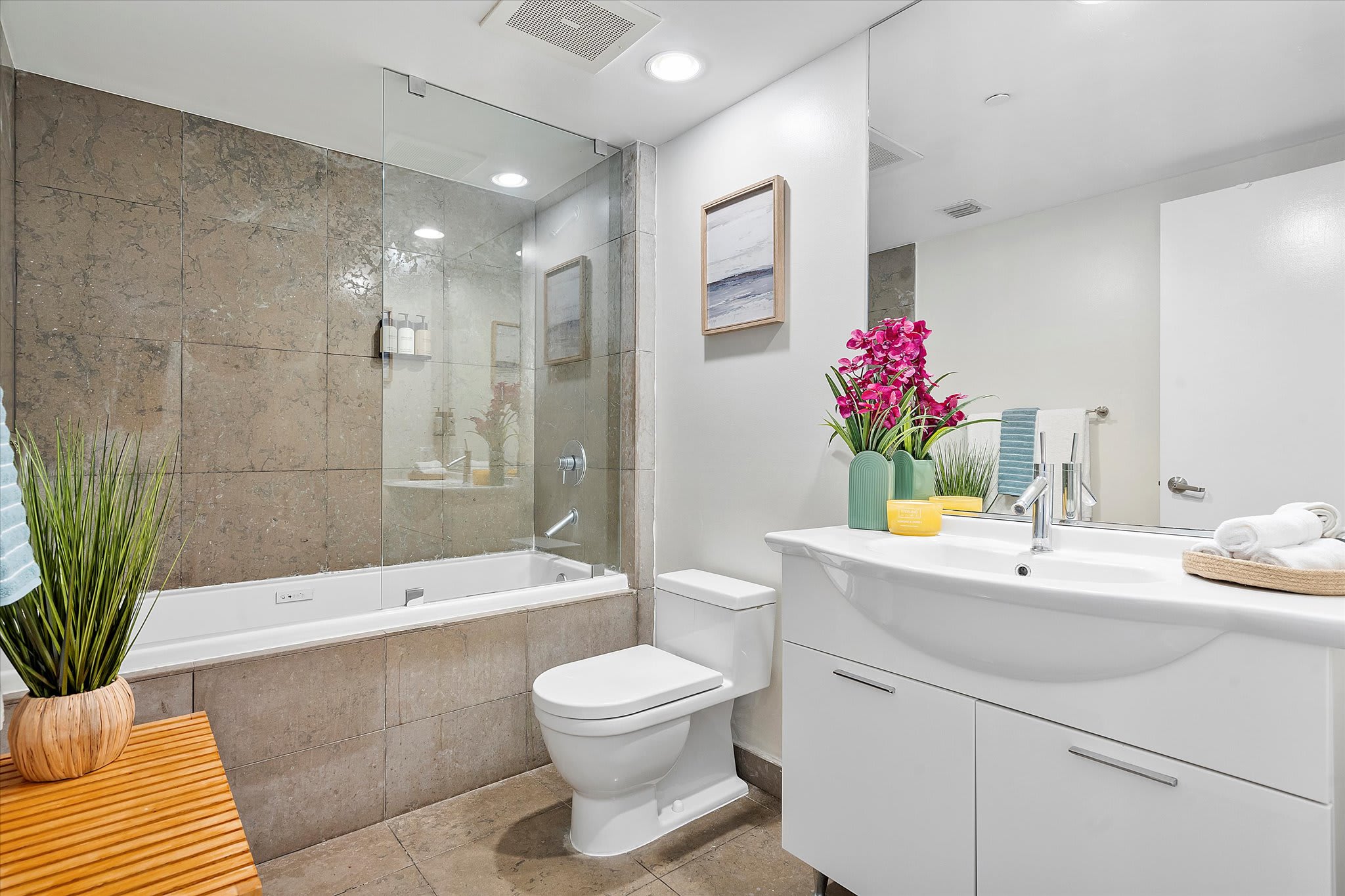 Clean and bright bathroom featuring a white vanity with a single sink and a large mirror, alongside a white toilet. The shower/tub combo with its grey-brown tiled walls and glass partition is visible, accented by vibrant pink flowers on the counter.