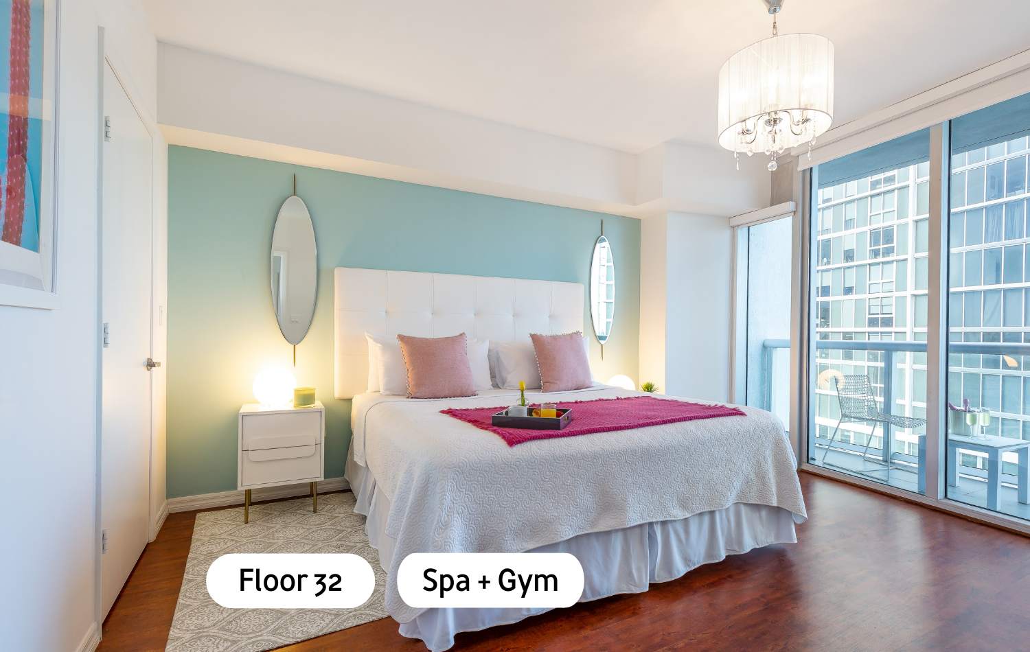 Bright bedroom with a white tufted headboard king bed, light blue accent wall, and a private balcony with city views. Two oval mirrors flank the bed, and a tray with breakfast items sits on a fuchsia throw.