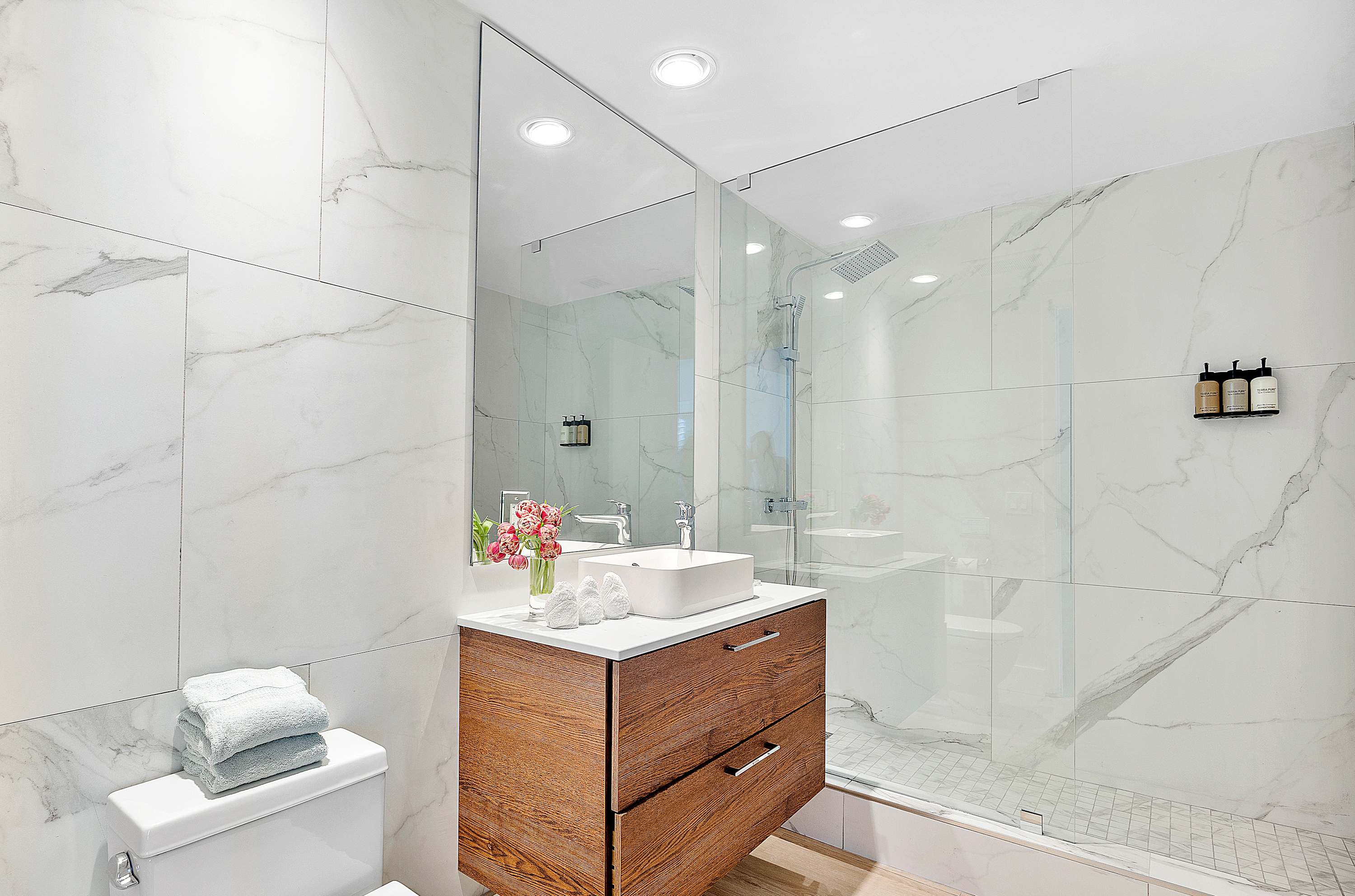 Modern bathroom featuring large white marble-patterned tiles on walls and shower, a floating wooden vanity with a white vessel sink, and a large frameless mirror. The glass-enclosed shower has a rain showerhead and built-in shelves.