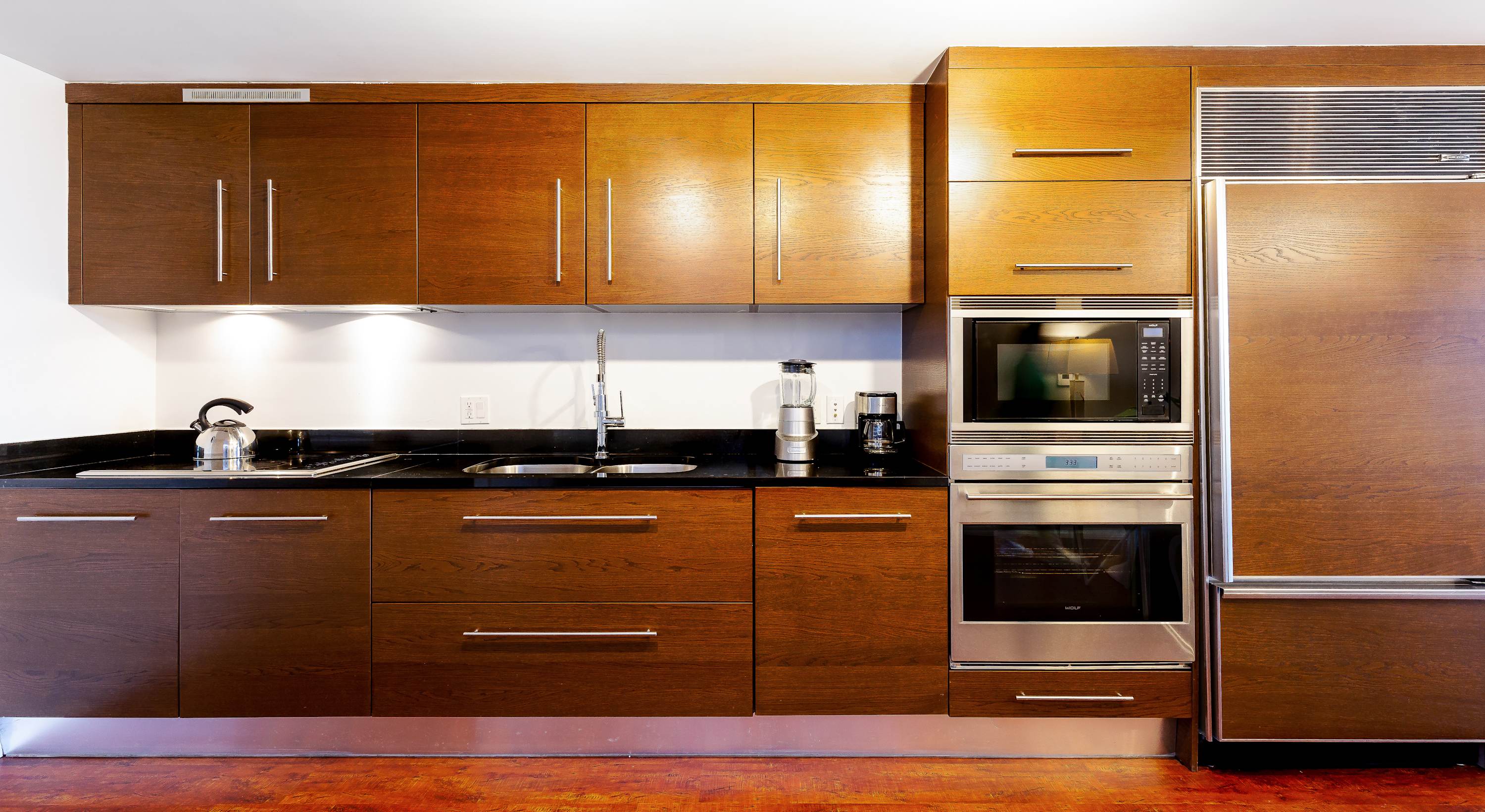 Modern kitchen with dark wood cabinetry, black granite countertops, and stainless steel appliances including a built-in oven, microwave, and refrigerator.