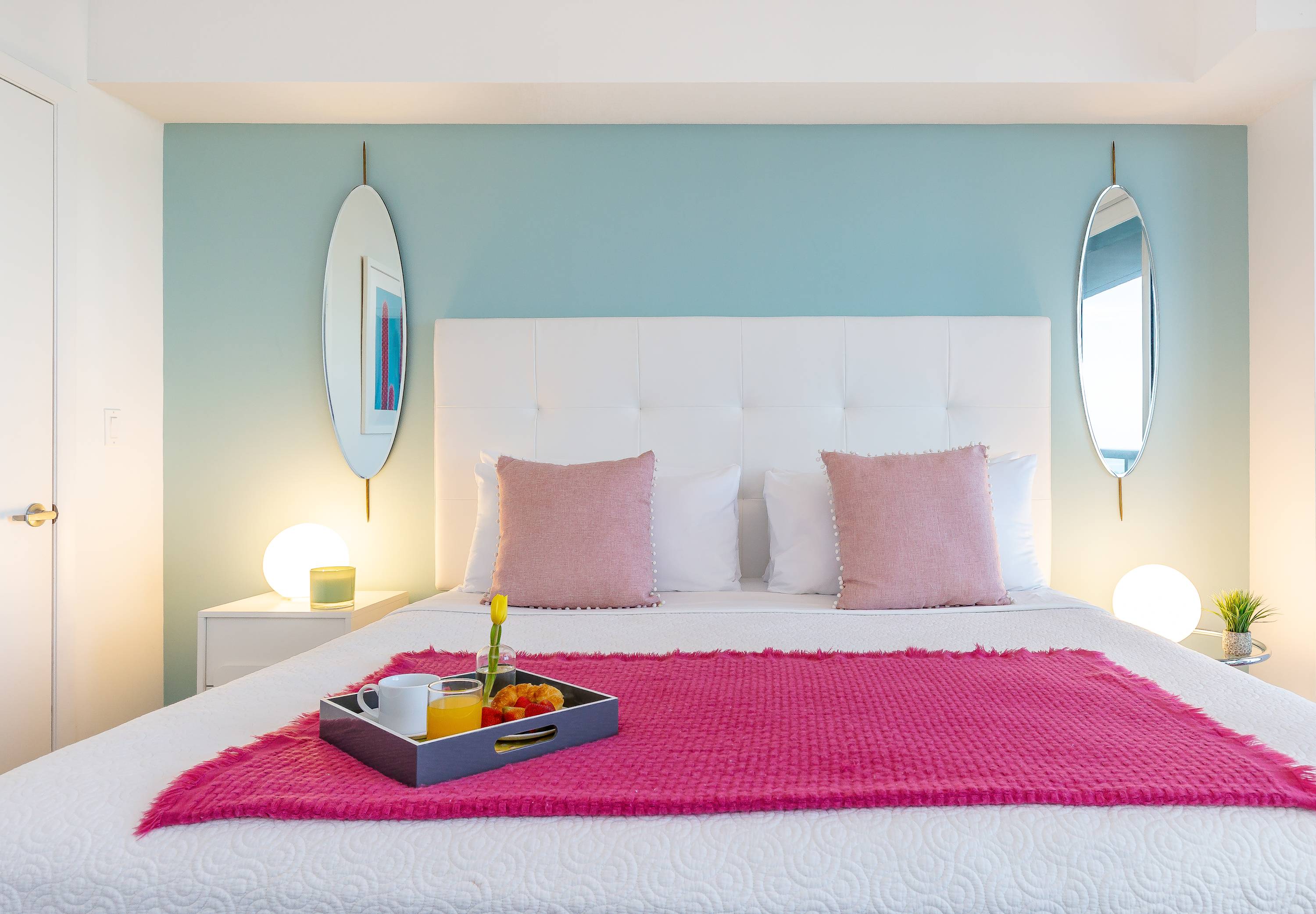 Close-up of a king-sized bed with a white tufted headboard, white bedding, and pink throw pillows. A fuchsia blanket and a breakfast tray with coffee and juice are placed on the bed.