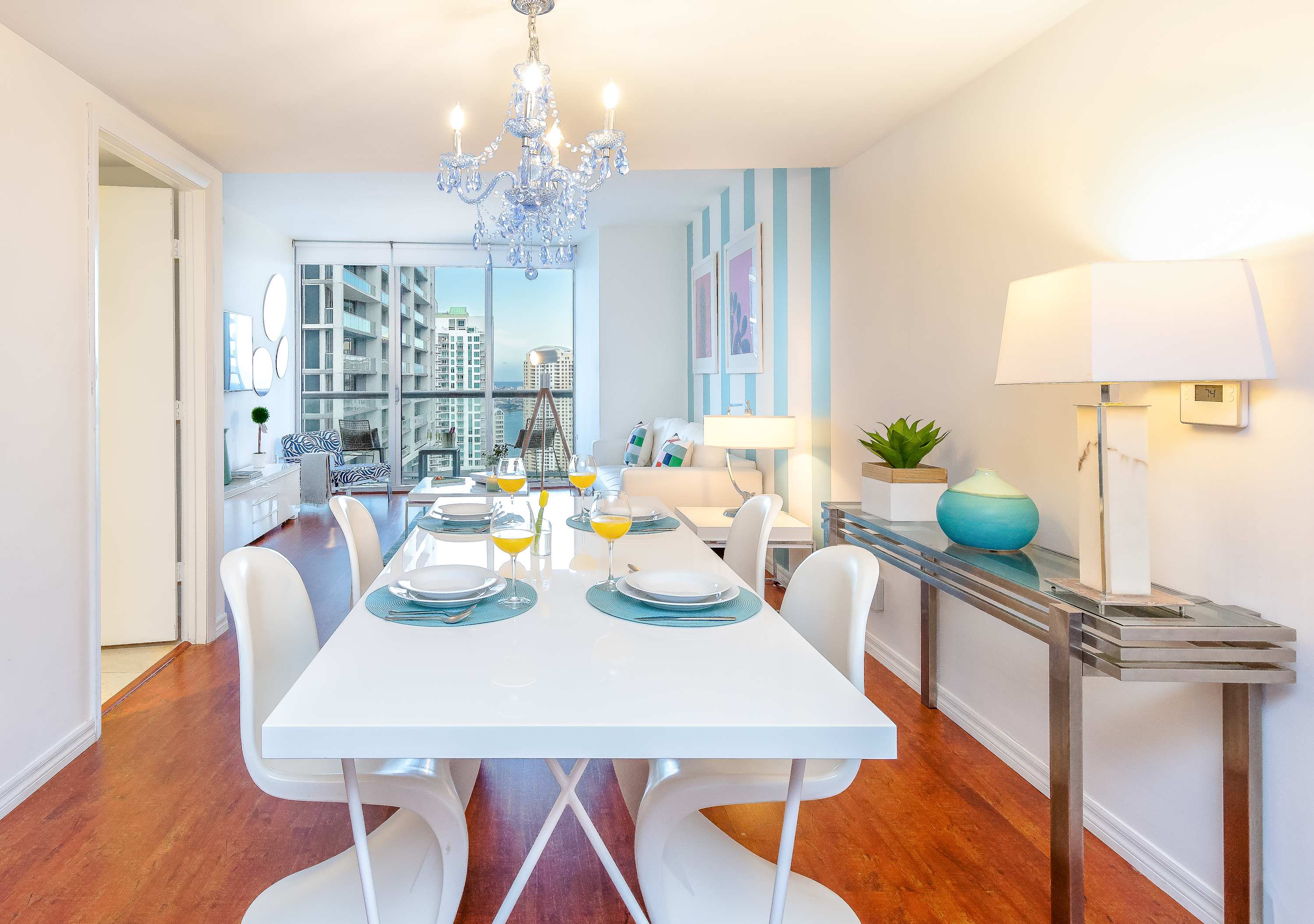 Modern dining area with a long white table and six white chairs, set with light blue placemats and yellow drinks. A blue chandelier hangs above, and a console table with a lamp is against the wall.