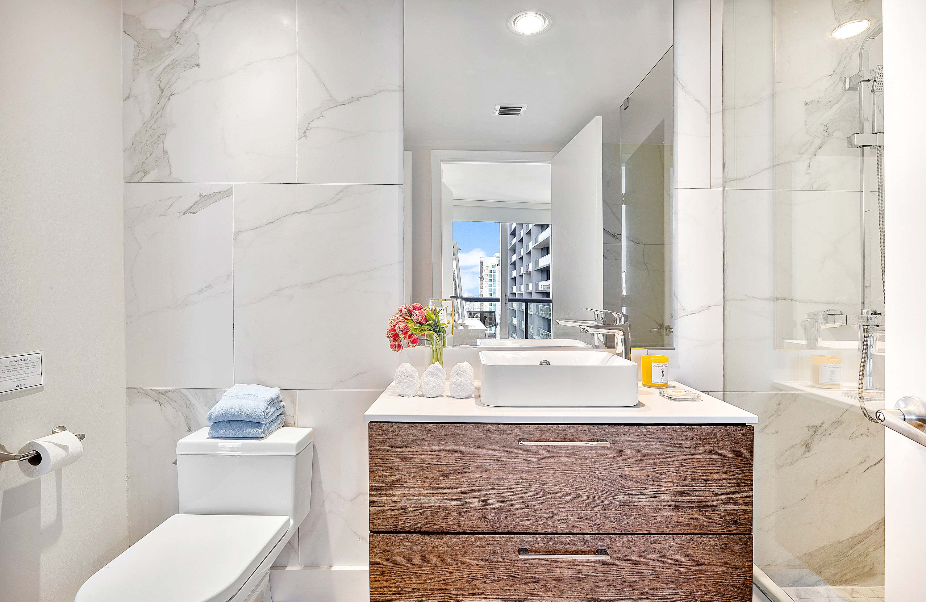 Modern bathroom with white marble-patterned tile walls, a floating dark wood vanity with a vessel sink, and a large mirror. A toilet is visible next to the vanity.