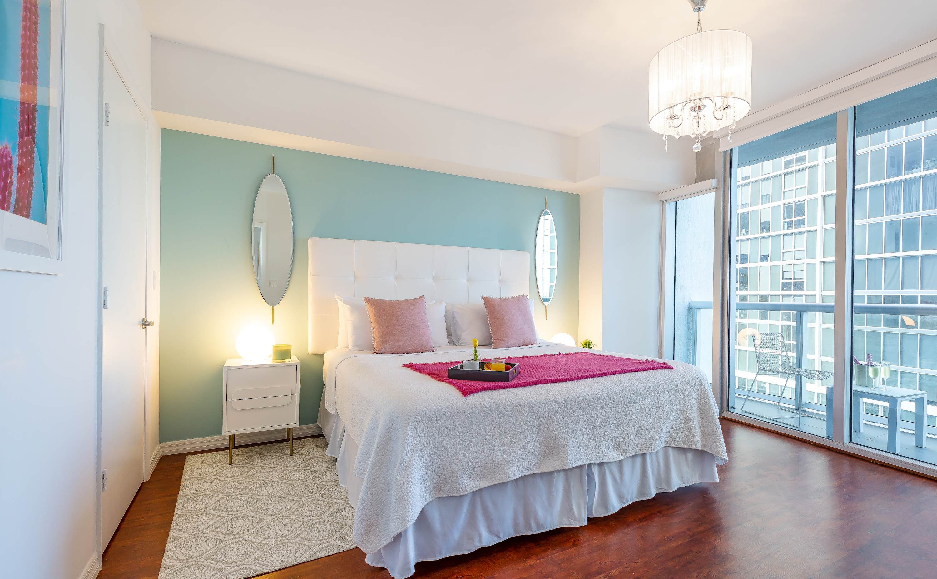 Bright bedroom with a white tufted headboard king bed, light blue accent wall, and a private balcony with city views. Two oval mirrors flank the bed, and a tray with breakfast items sits on a fuchsia throw.