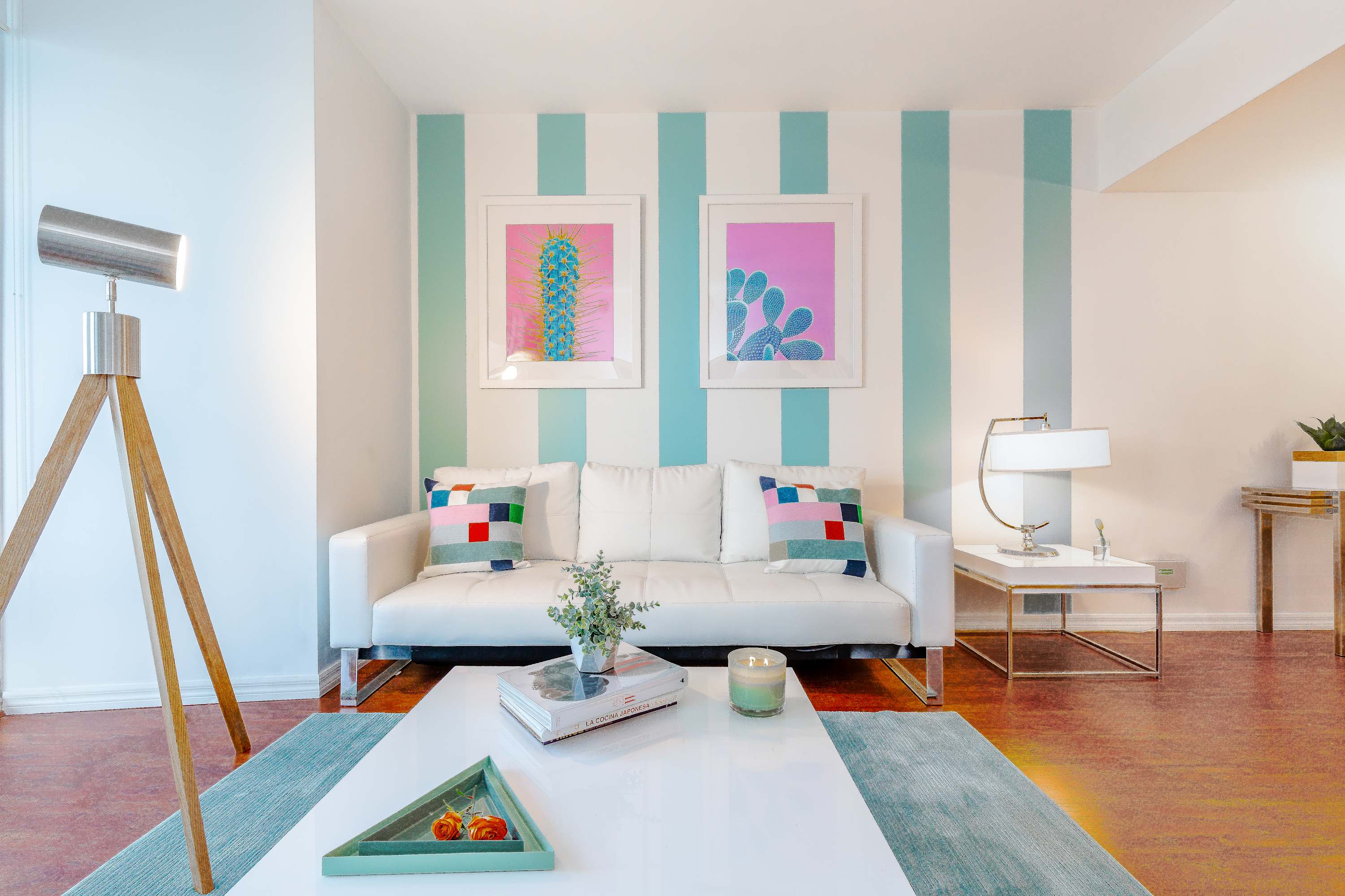 Cozy living room with a white sofa, colorful throw pillows, and a white coffee table on a teal rug. A striped accent wall features two framed cactus prints, and a modern floor lamp stands nearby.