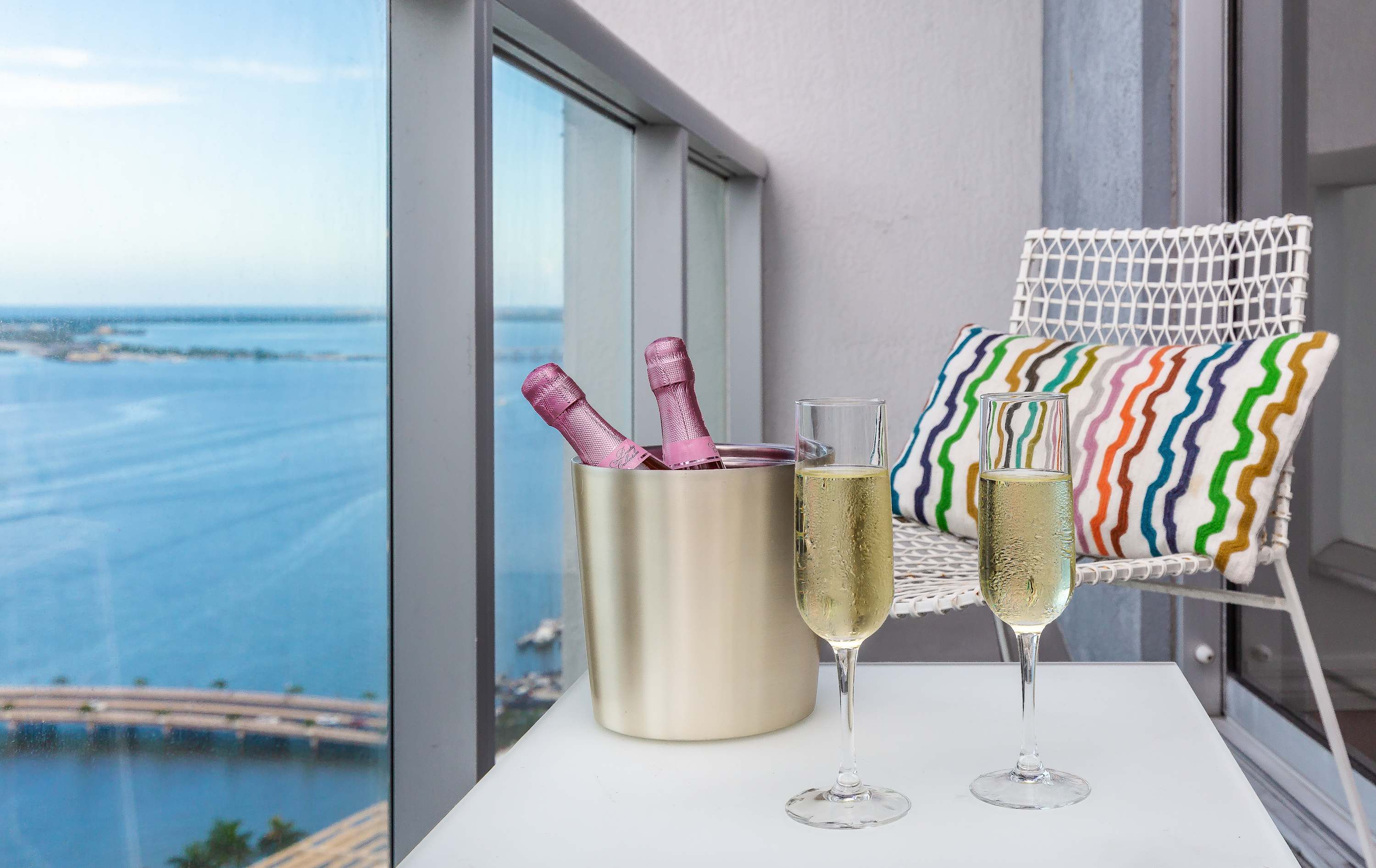 Balcony with a stunning waterfront view, featuring a small white table with two champagne flutes and a bucket with two bottles of rosé champagne. A white woven chair with a colorful striped pillow is visible.