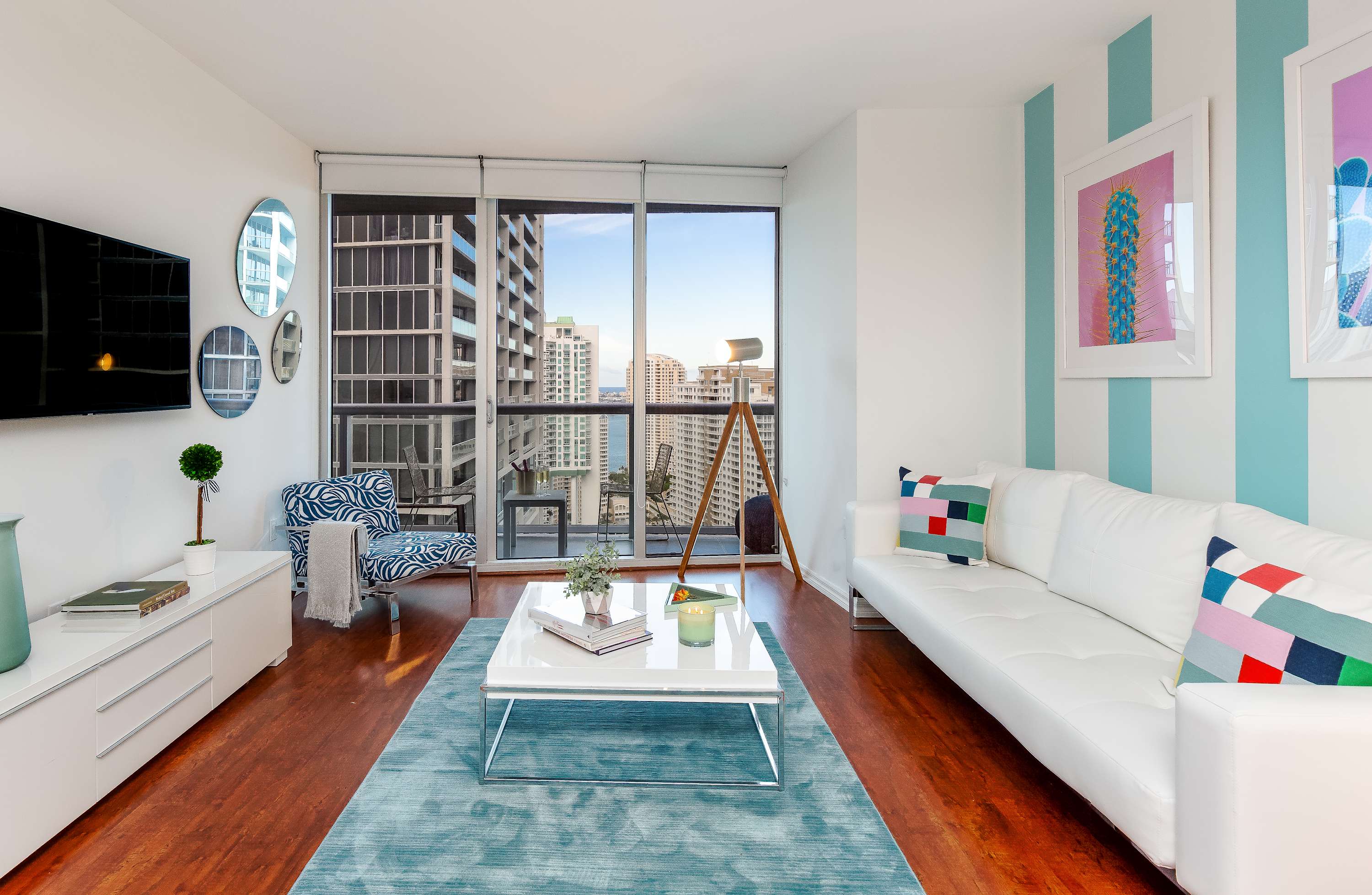 Contemporary living room featuring a white sofa with colorful throw pillows, a white coffee table on a teal rug, and a zebra-print armchair. Floor-to-ceiling windows offer expansive city views and access to a balcony.