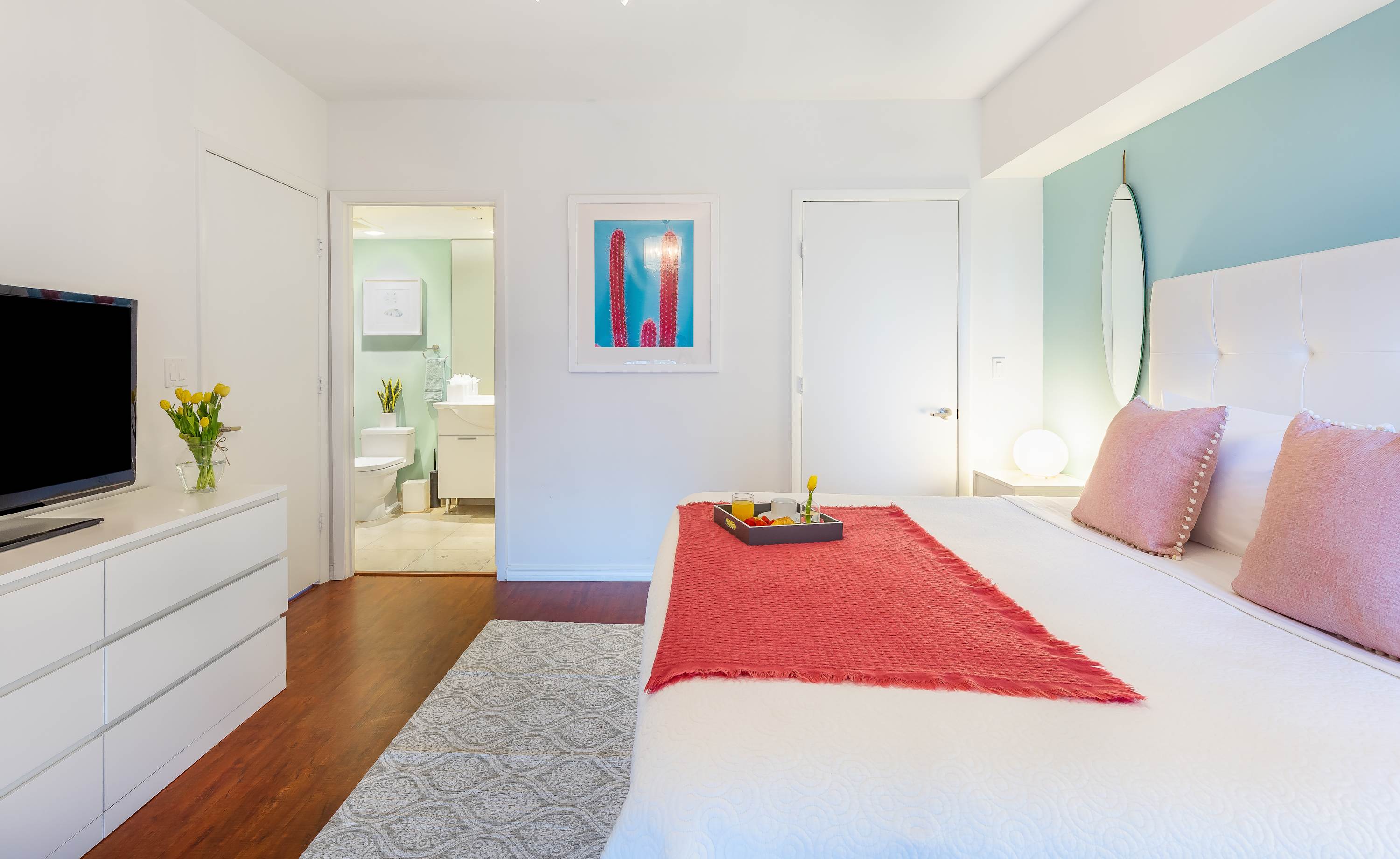 King bedroom with a white tufted headboard, light blue accent wall, and a white dresser with a TV. An open doorway leads to a modern bathroom, and a framed cactus print hangs on the wall.