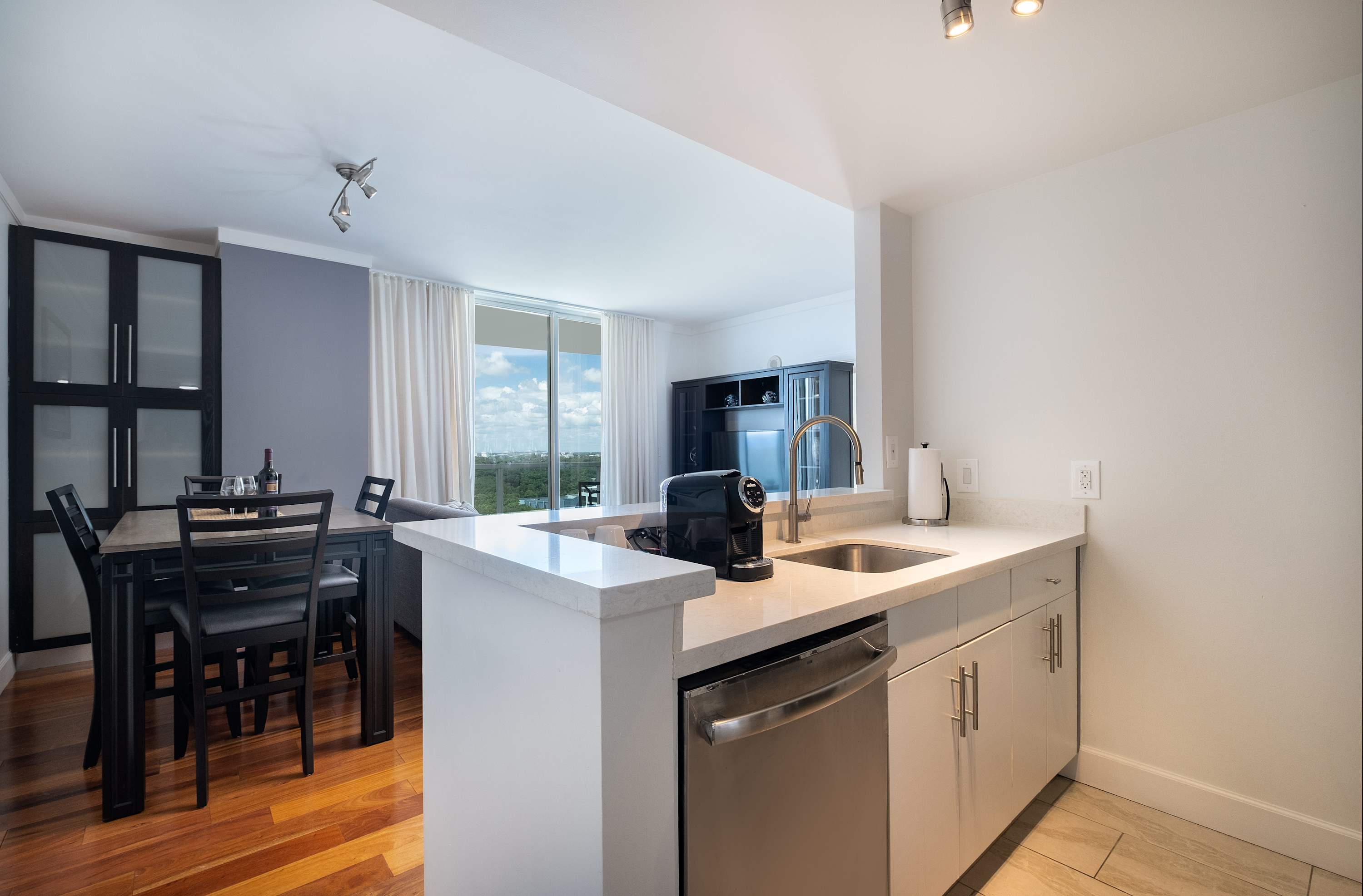 Modern kitchen with white quartz countertops, stainless steel appliances including a dishwasher and sink, and light wood flooring. A dining area with a dark wood table and chairs is visible in the background.