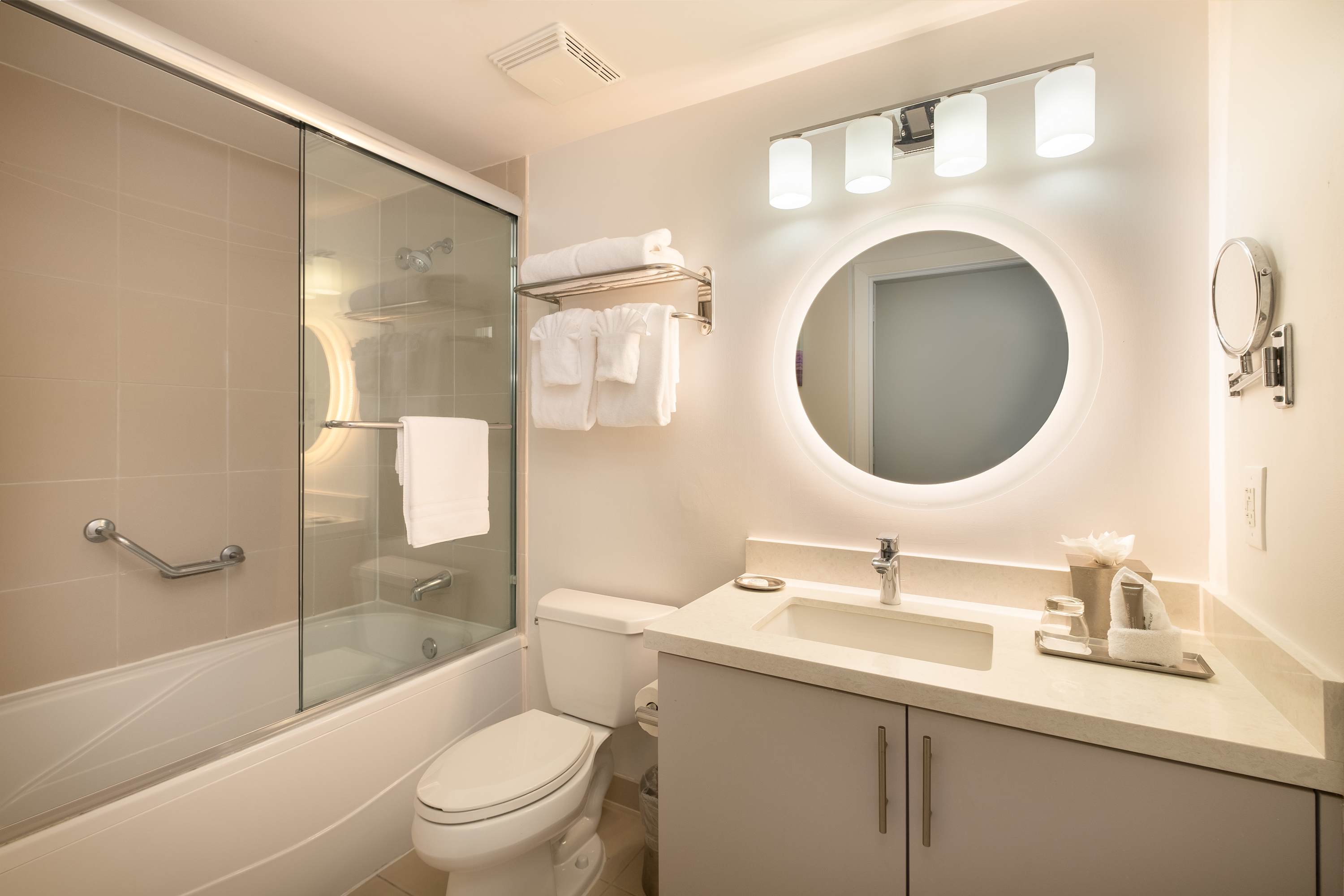 Bright bathroom with a white vanity, a round backlit mirror, and a shower-over-tub with a glass enclosure. White towels are neatly stacked on a rack, and the room features light-colored tiles.