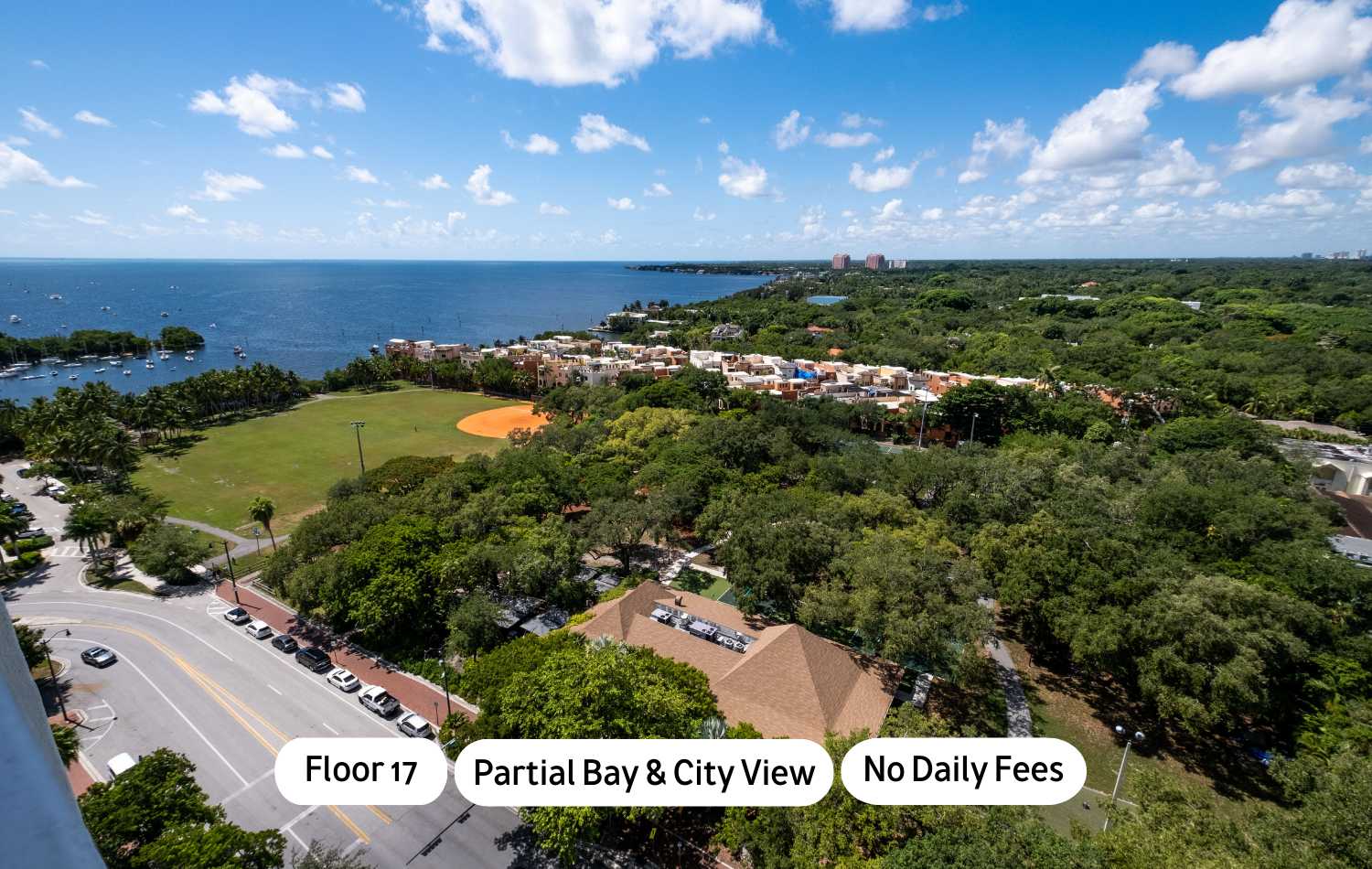 High-angle view showcasing a vast bay with numerous boats, a baseball field, lush green trees, and distant city buildings under a bright blue sky with white clouds. Text overlays indicate 'Floor 17', 'Partial Bay & City View', and 'No Daily Fees'.