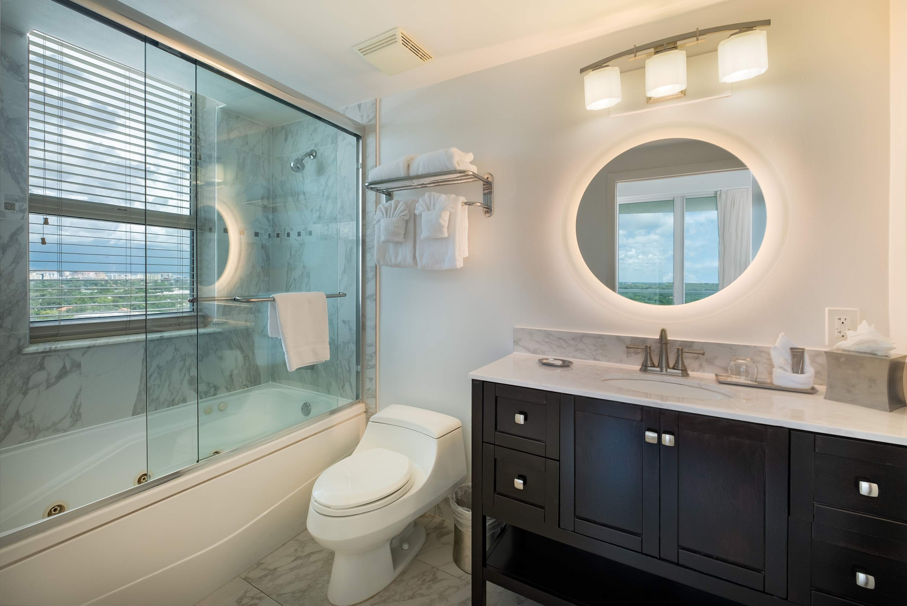 Elegant bathroom featuring a dark wood vanity with a marble countertop, a round backlit mirror, and a shower-over-tub with a glass enclosure and marble-tiled walls. A window with blinds offers natural light.