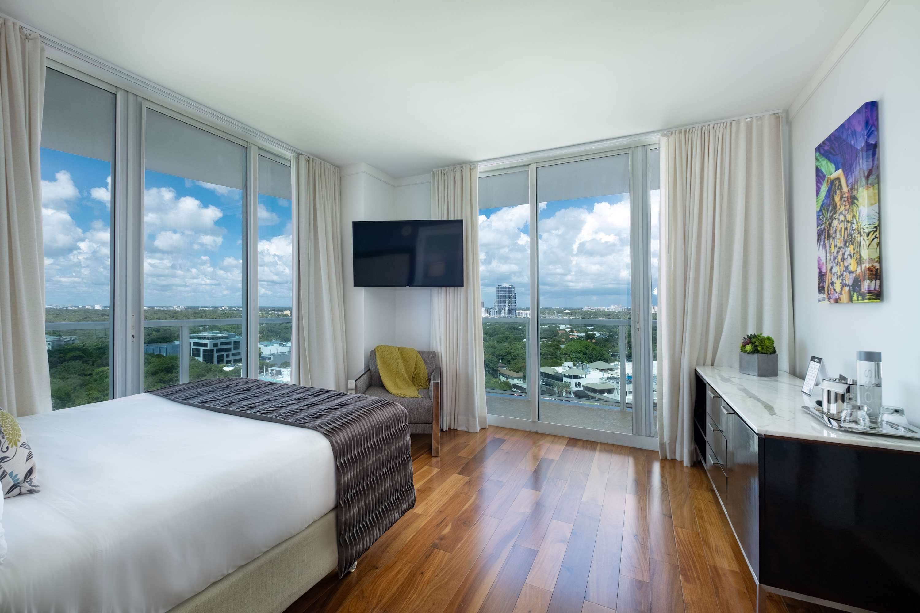 Bright bedroom featuring a large bed with a grey and white patterned throw, light hardwood floors, and expansive floor-to-ceiling windows offering panoramic city and partial bay views. A wall-mounted TV and a modern armchair complete the contemporary decor.
