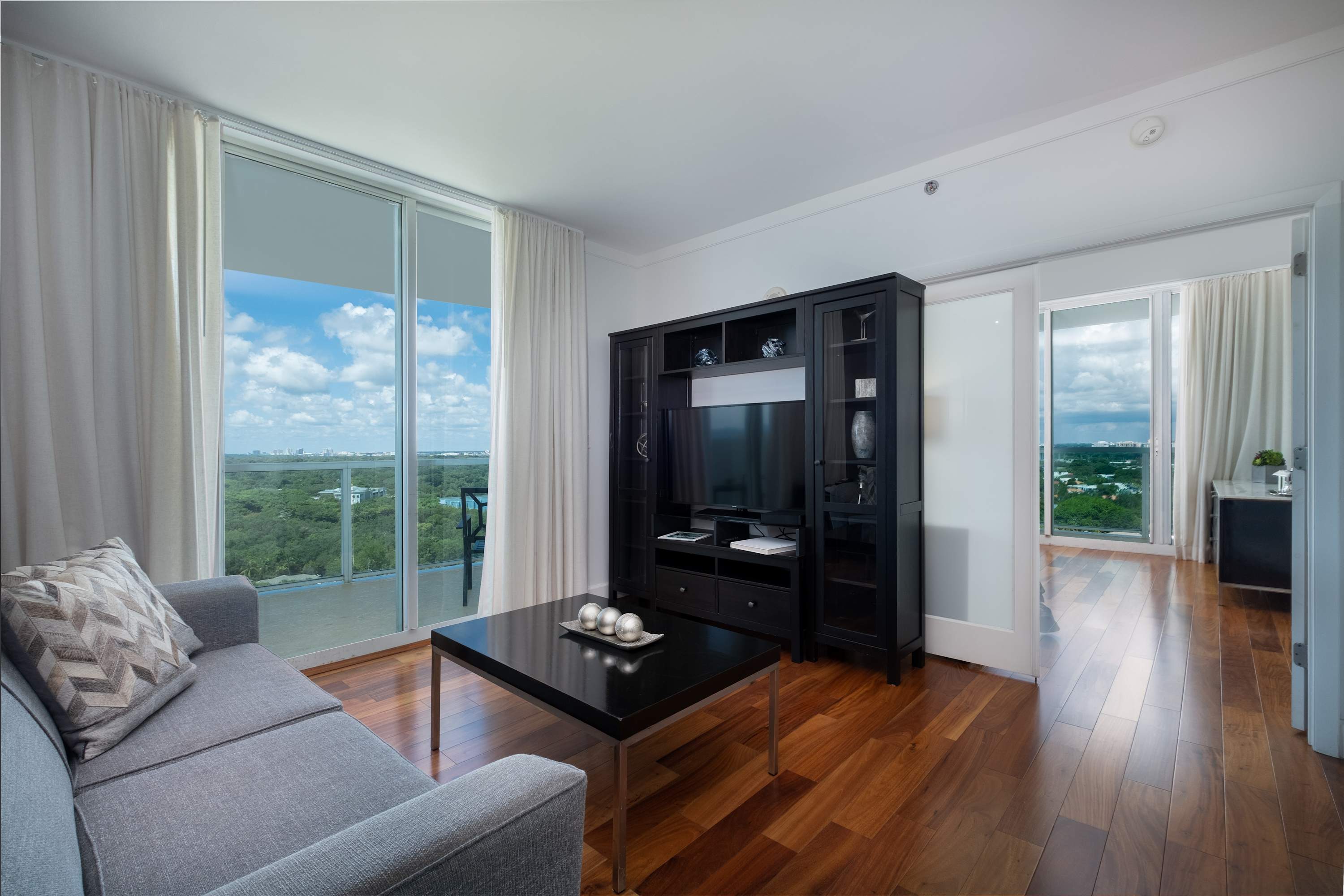 Spacious living room with a grey sofa, dark coffee table, and a large black entertainment unit housing a TV, all set on polished hardwood floors. Expansive sliding glass doors open to a balcony with city views.