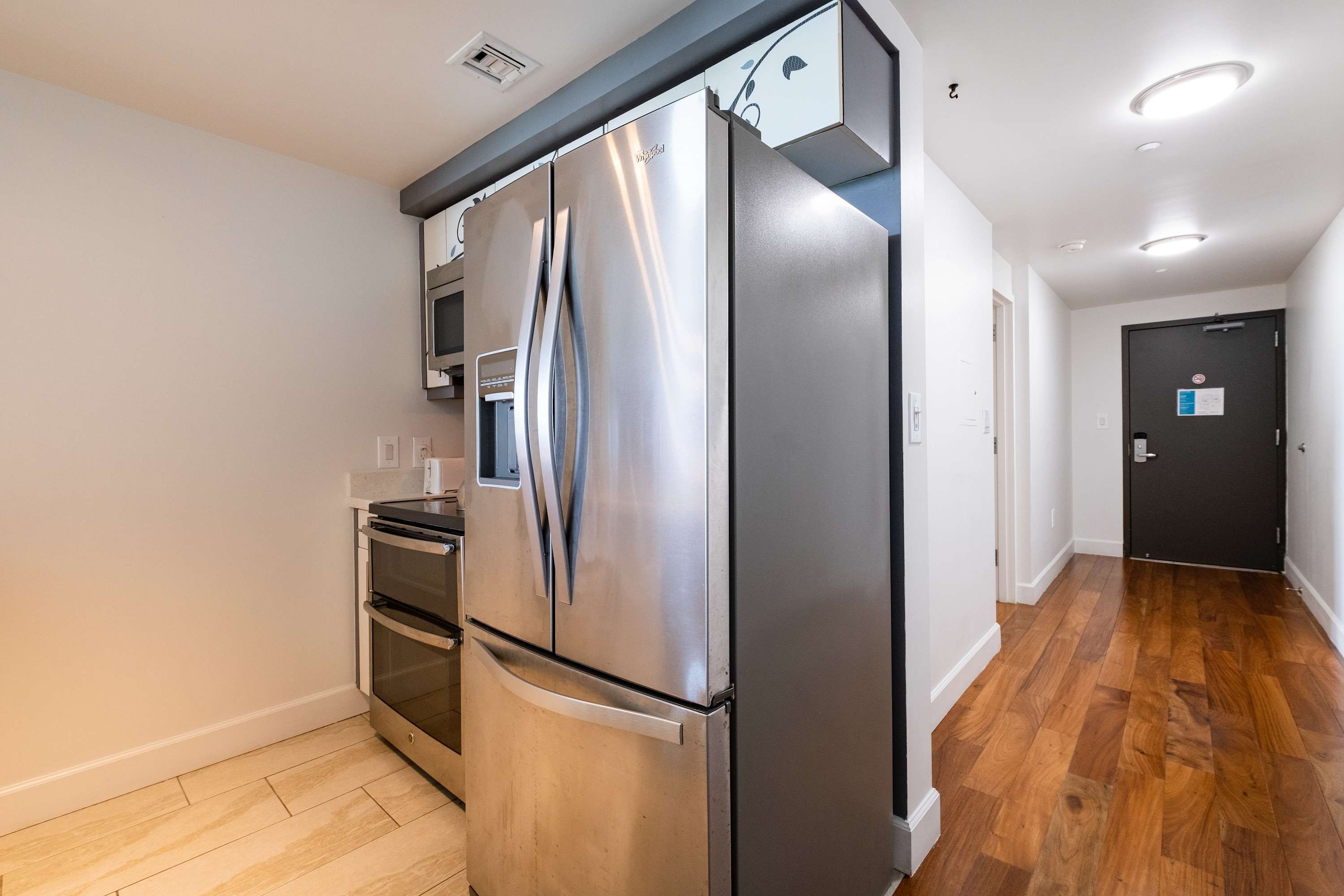 Compact kitchen area featuring a large stainless steel refrigerator, a microwave, and a small range, set against light-colored walls and tiled flooring. A hallway with hardwood floors leads to an entrance door.