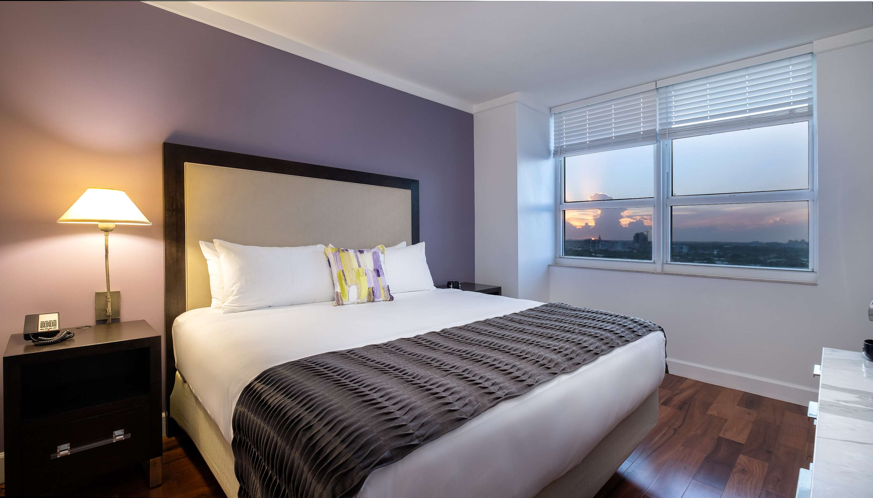 Cozy bedroom featuring a large bed with a grey and white patterned throw, a purple accent wall, and light hardwood floors. A window with blinds offers a partial city view, and a bedside lamp provides warm lighting.