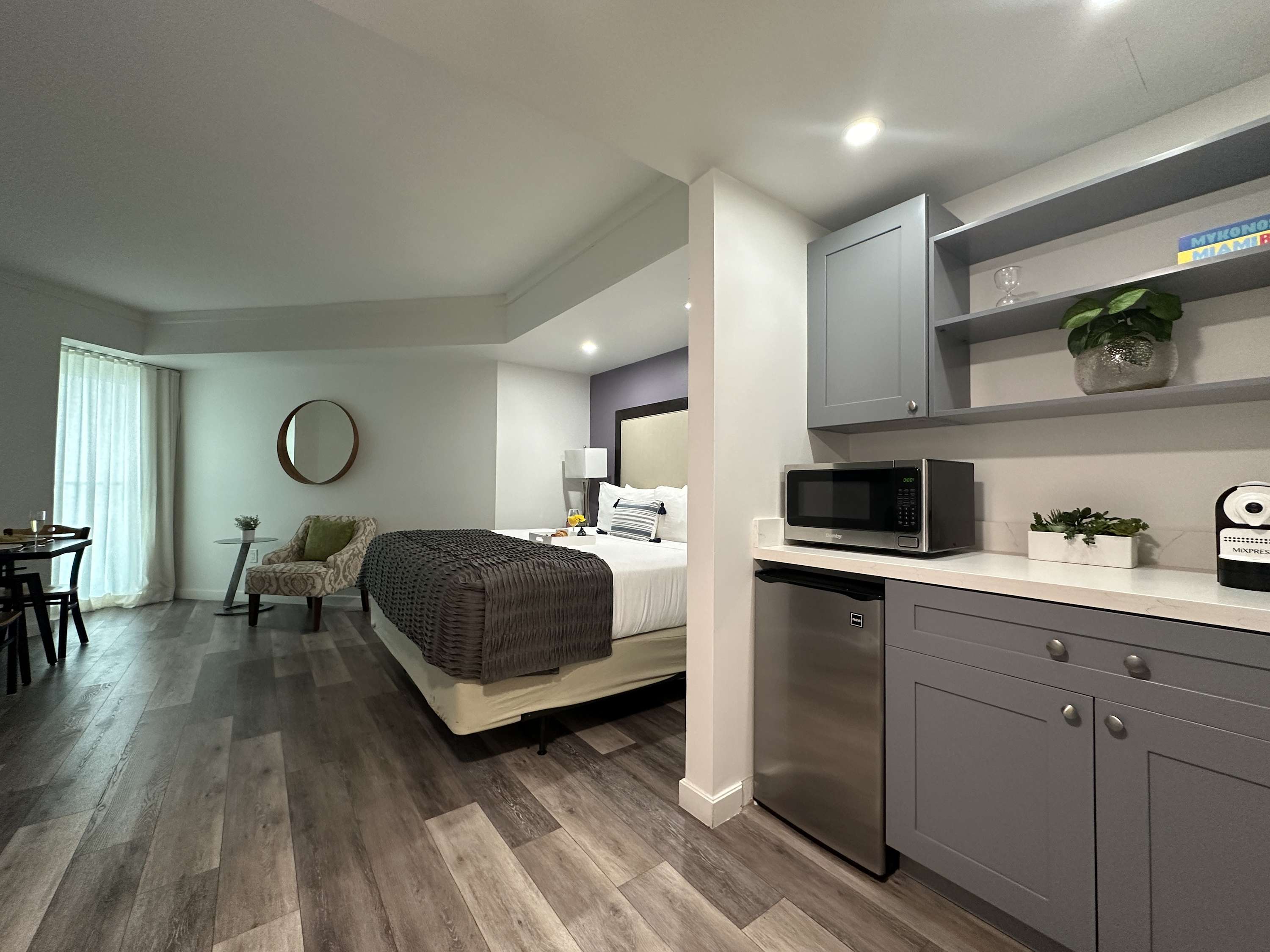 Well-appointed studio room showcasing a compact kitchenette with gray cabinetry, a microwave, and a mini-fridge, adjacent to a comfortable bed with a purple accent wall and light wood-look flooring.