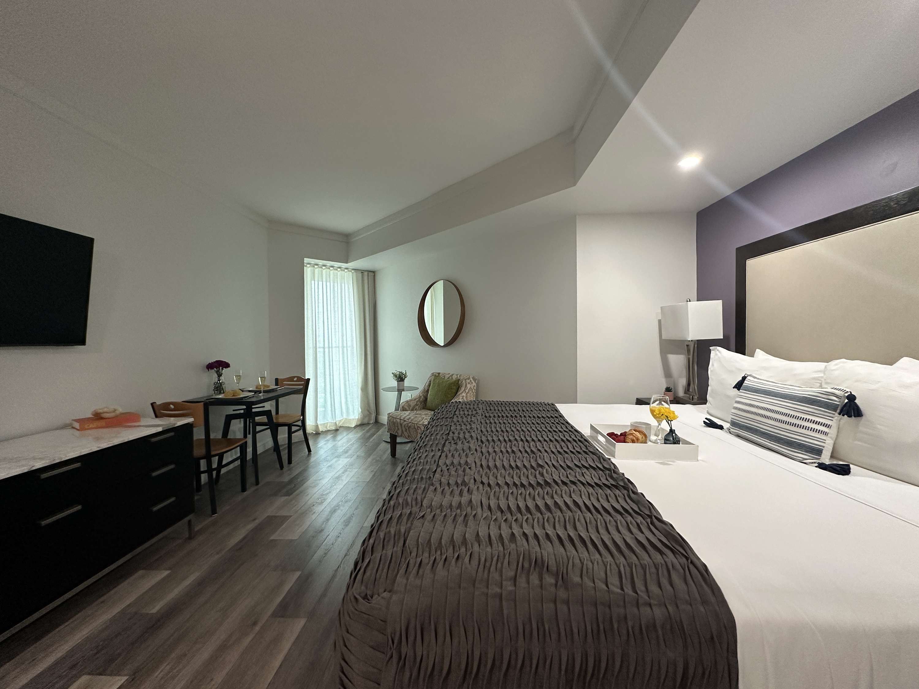 Inviting studio space with a large bed, a small dining table for two, and a long console table with a TV, all illuminated by natural light from a large window and recessed lighting.