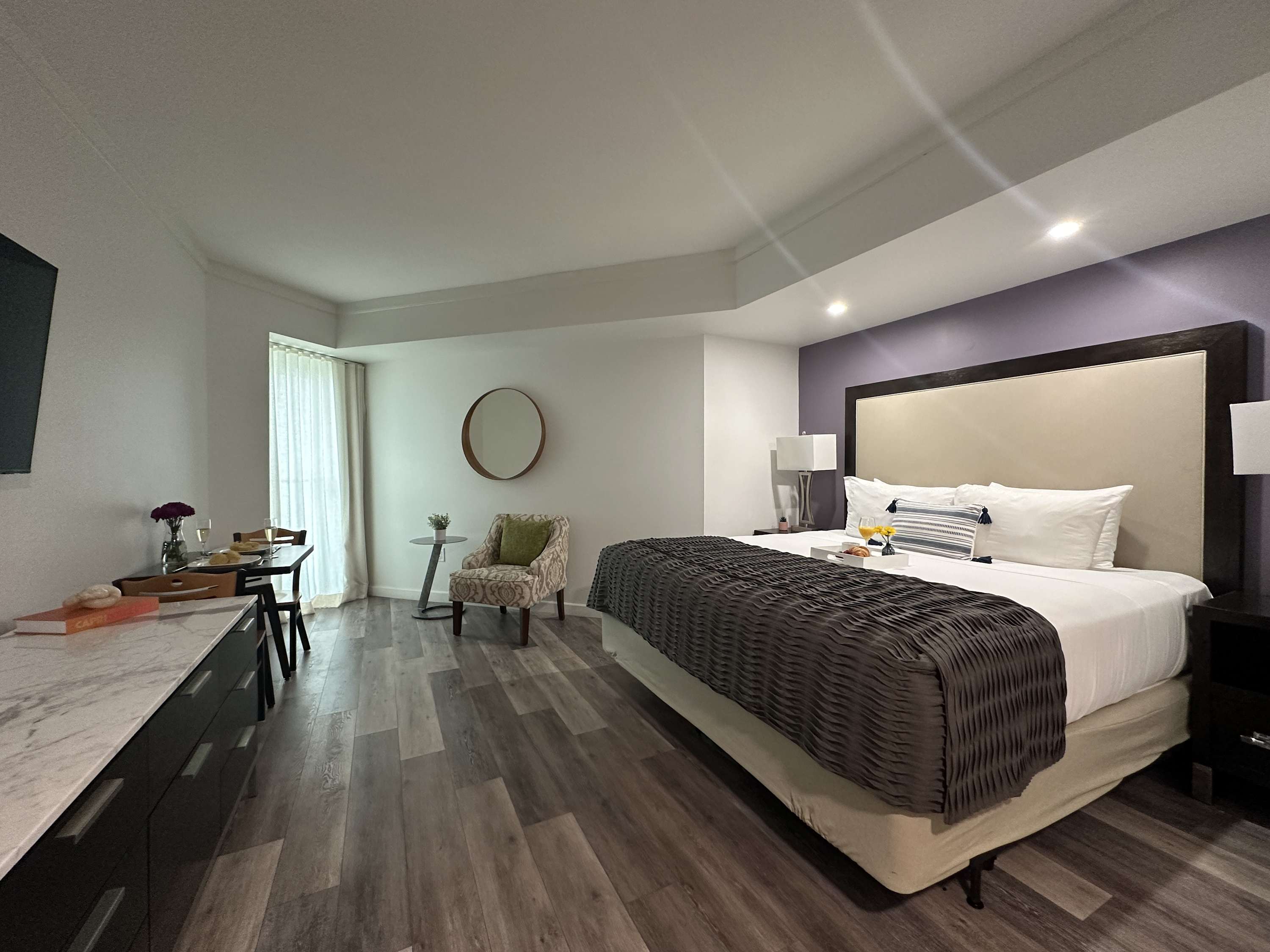 Expansive studio room with a large bed, a small dining table with chairs, a wall-mounted TV, and a comfortable armchair, all set on light wood-look flooring with a large window.