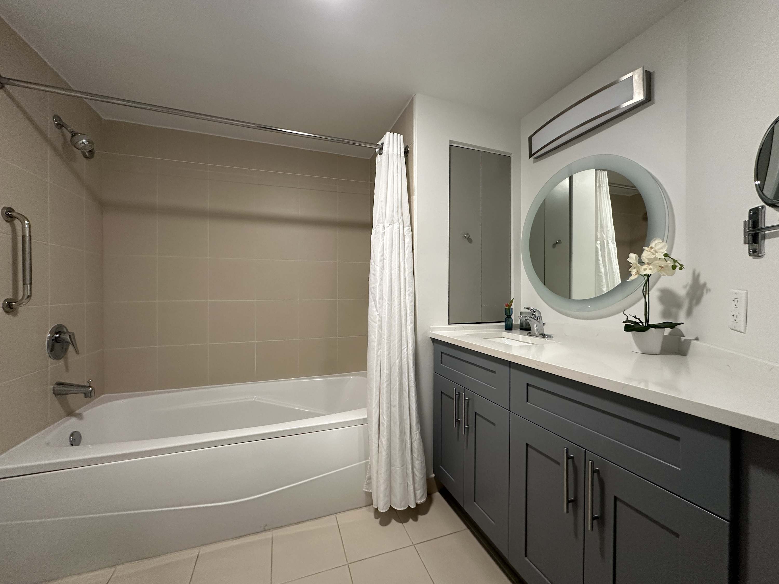 Bright bathroom featuring a white bathtub with a shower curtain, light beige tiled walls, and a modern vanity with gray cabinets and a round illuminated mirror.