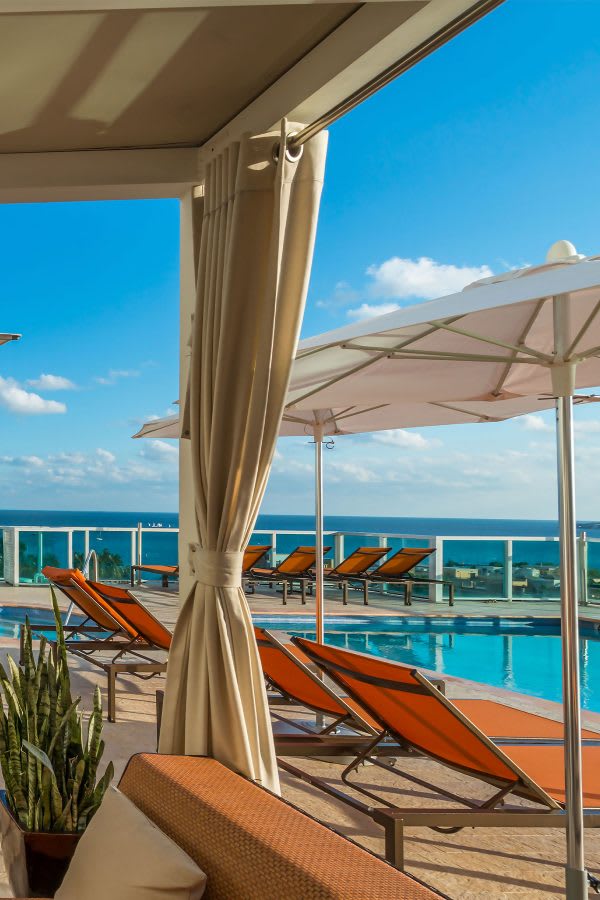 Sunny outdoor pool deck featuring orange lounge chairs, white umbrellas, and a shaded cabana area with flowing curtains, all set against a bright blue sky and distant ocean. A green plant adds a touch of nature.
