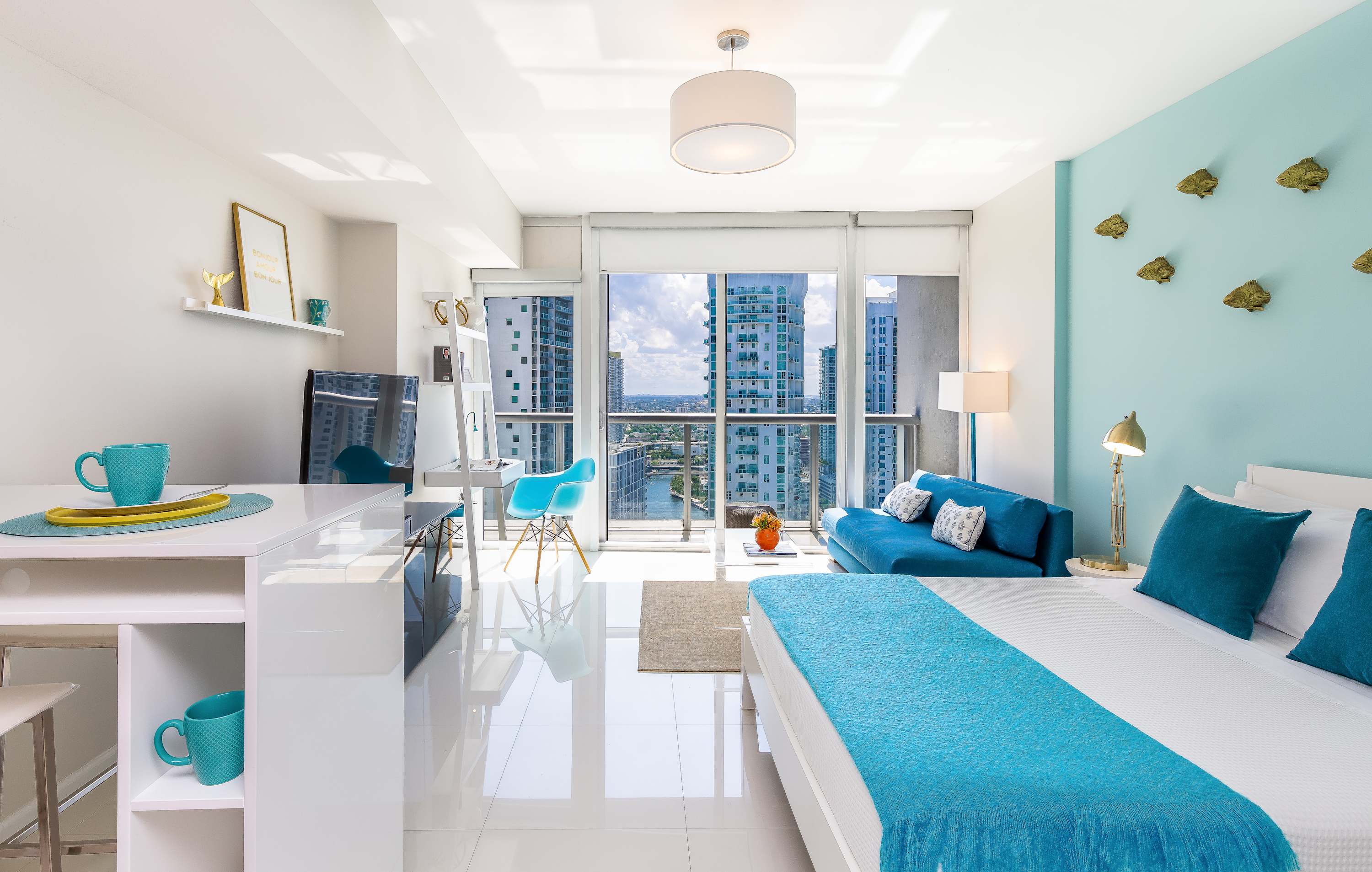 Open-concept studio apartment featuring a bed with a blue throw, a blue sofa, a white dining/desk area, and a compact kitchen, all with white tiled floors and large windows overlooking the city.
