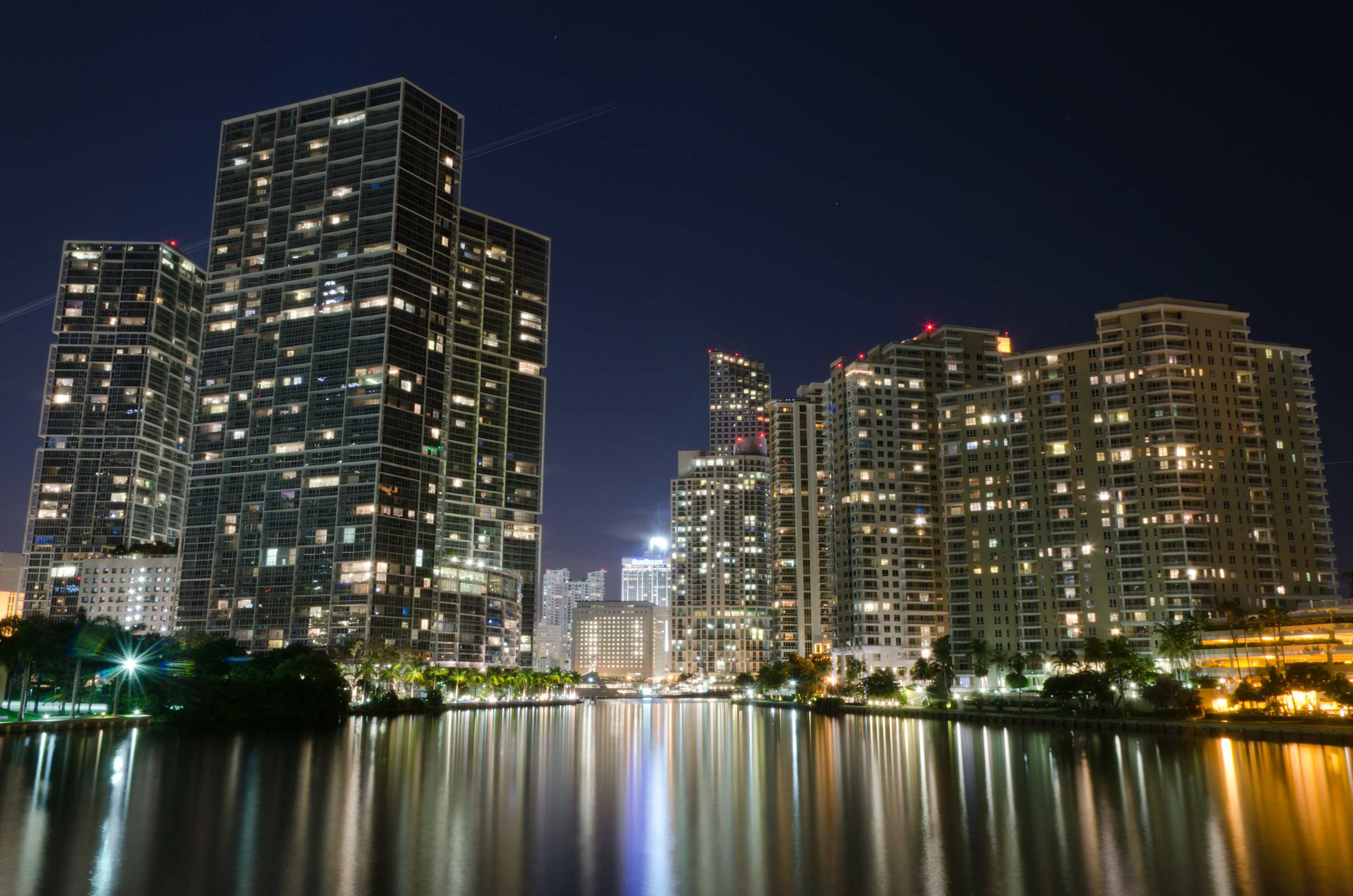 A stunning nighttime cityscape showcases numerous illuminated high-rise buildings reflecting their lights on a tranquil body of water, creating a vibrant and dramatic urban panorama under a dark sky.