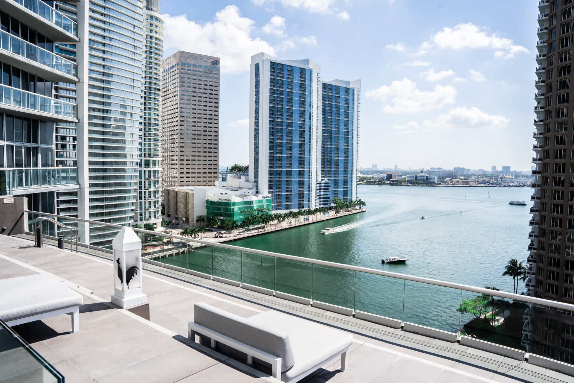 A spacious balcony offers panoramic waterfront views of Miami's urban landscape, featuring multiple modern high-rise buildings and boats navigating the calm bay. Two contemporary outdoor lounge chairs are positioned for relaxation.