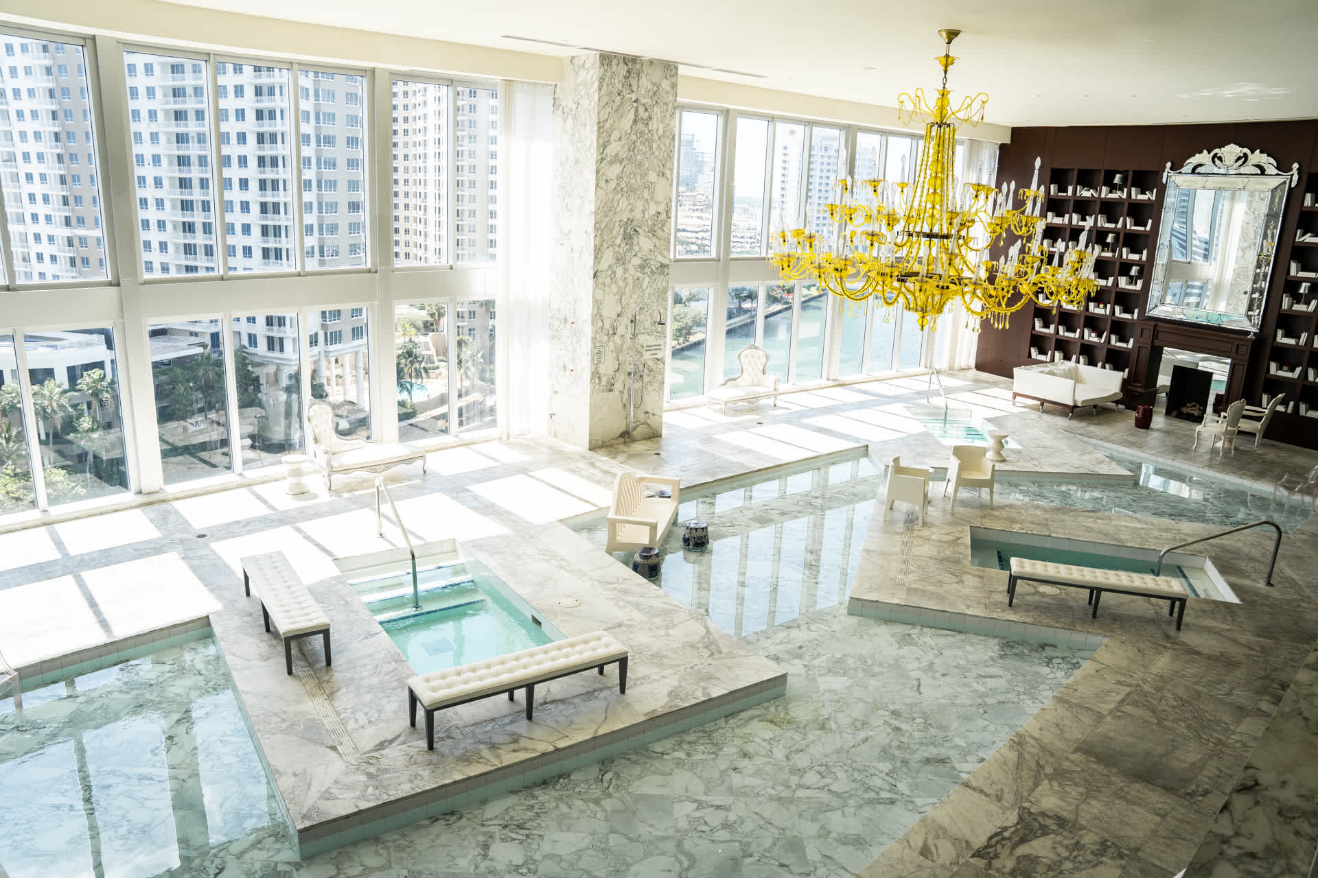 Luxurious indoor spa area featuring multiple shallow pools with marble surrounds, white benches, and a grand gold chandelier. Floor-to-ceiling windows offer panoramic city and river views.
