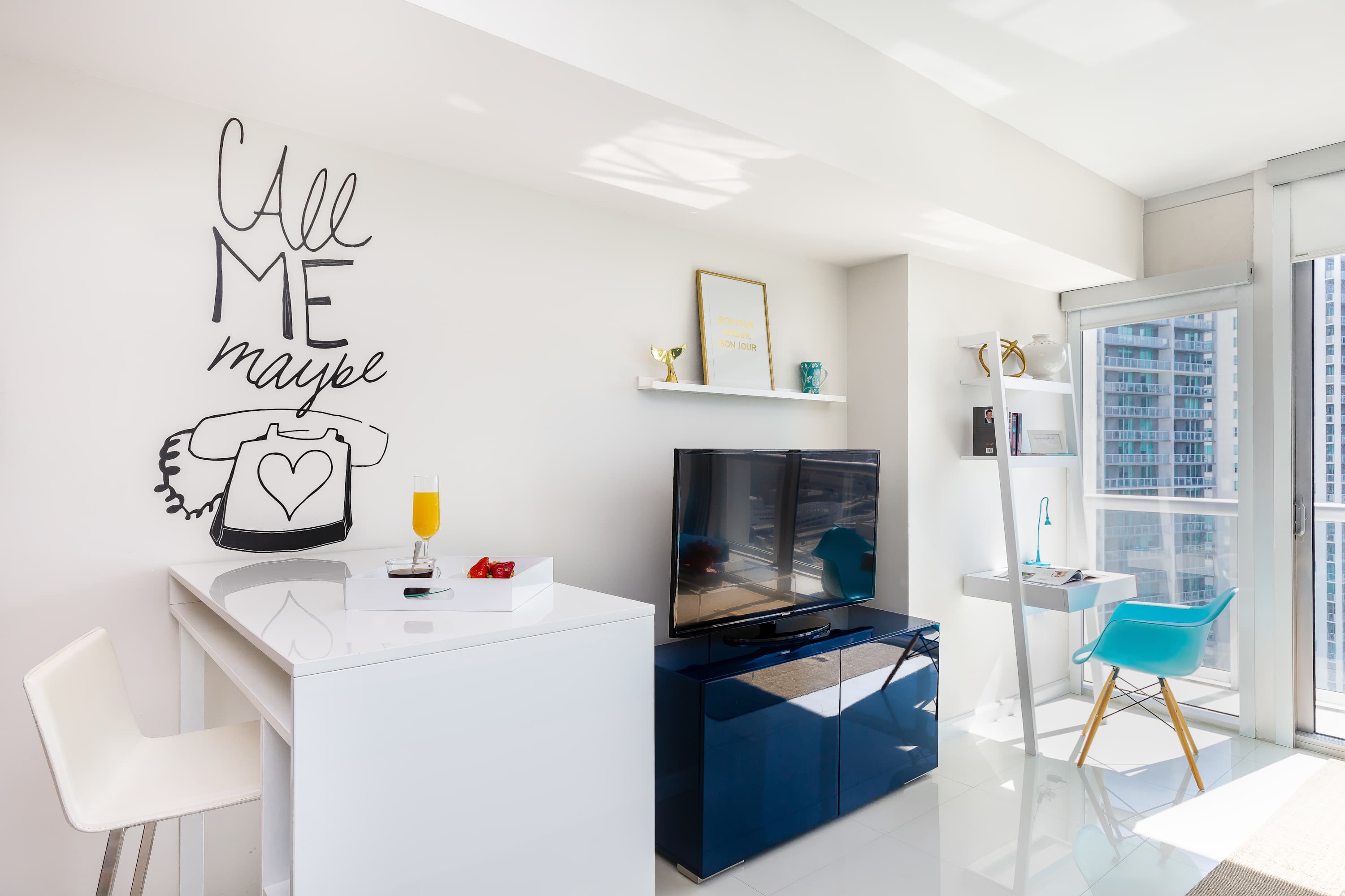 Bright living area with a white dining/desk counter, a wall-mounted TV on a blue console, and a white ladder shelf with a desk, all under a playful "Call Me Maybe" wall art.