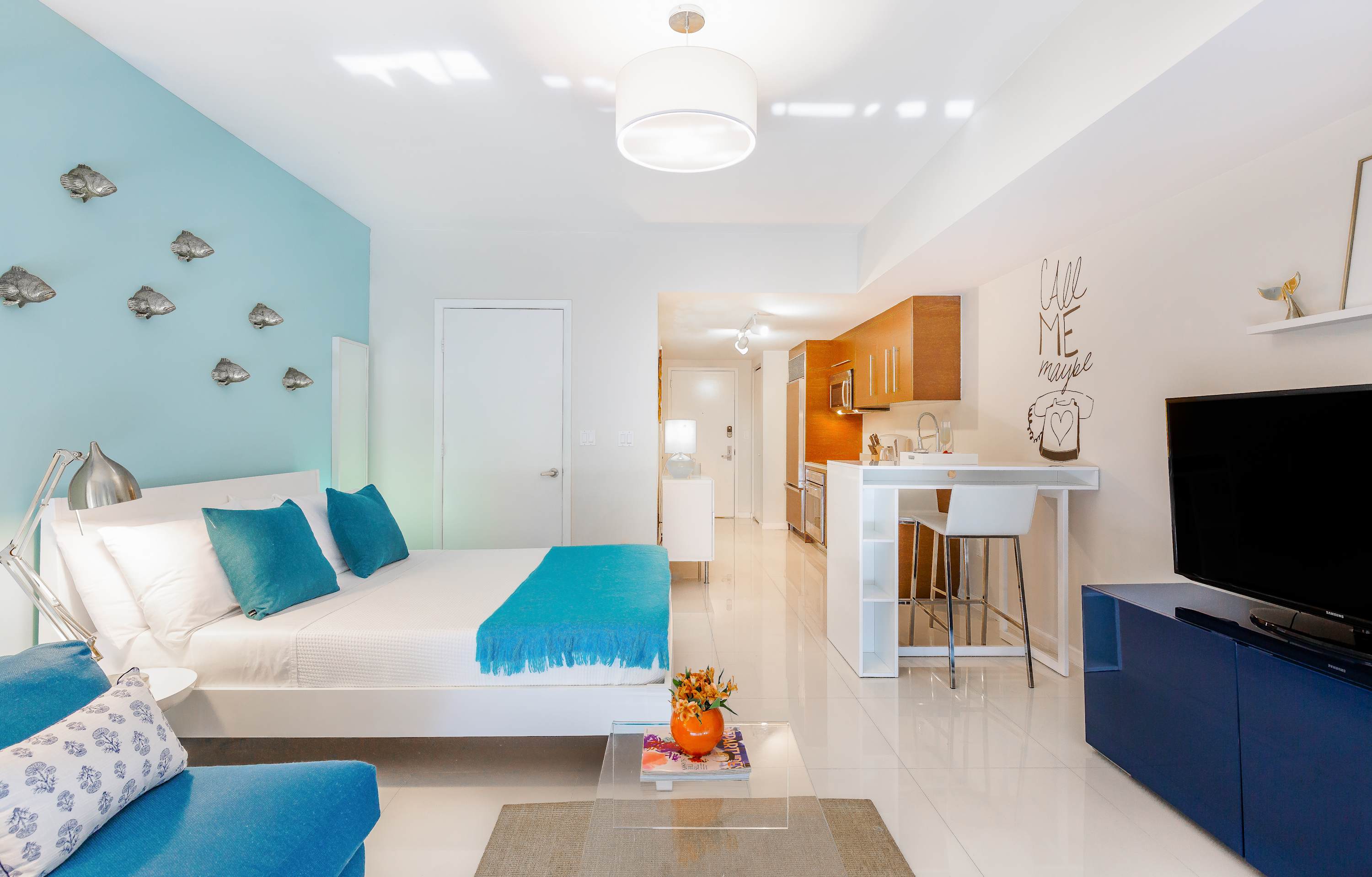 Wide shot of the studio apartment, showcasing the bed with blue accents, a blue sofa, a white dining counter, and a modern kitchen with wooden cabinets, all on glossy white floors.