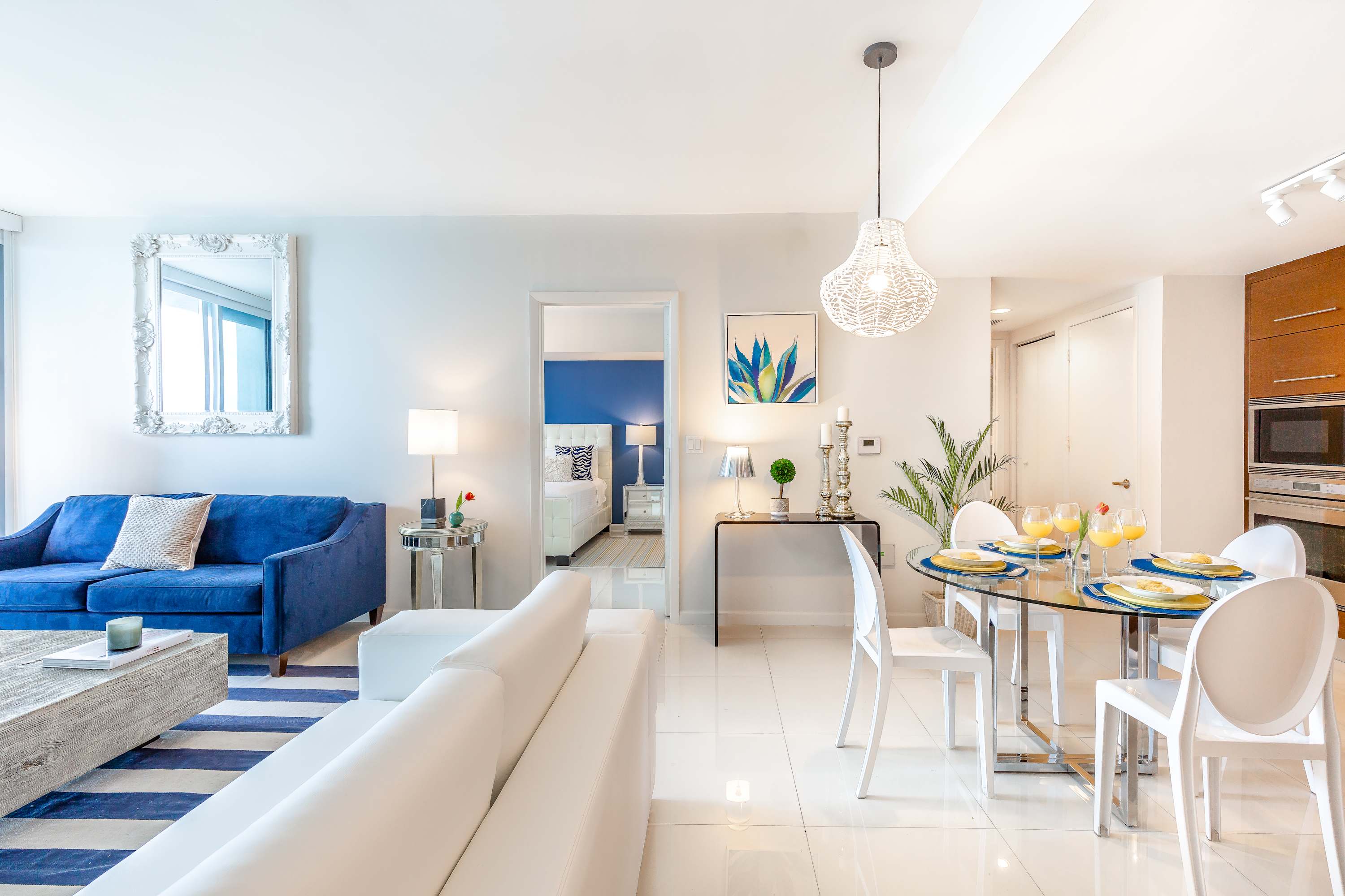 An open-concept living and dining space with white tile floors, a blue sofa, a white sofa, and a glass dining table with white chairs. A large ornate mirror hangs on the wall, and a glimpse of the bedroom with a blue accent wall is visible through a doorway.